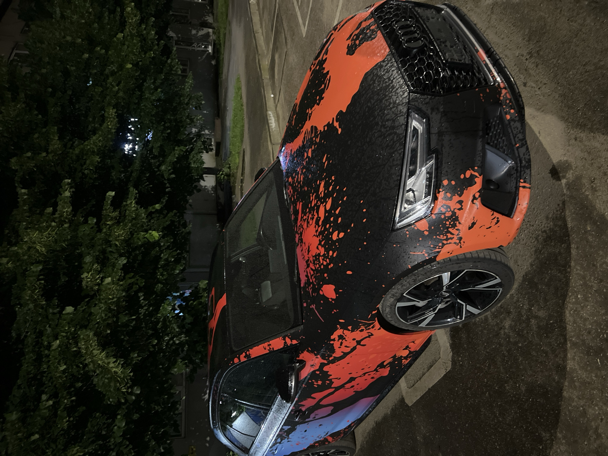 This vertical image, likely captured at night in Krasnodar, Russia, prominently features a custom-designed Audi car. The car, identifiable by its distinctive Audi four-ring logo on the honeycomb grille and modern LED headlights, sports a unique aesthetic. Its body is predominantly black, overlaid with an eye-catching, vivid orange splatter-paint or wrap pattern that covers the hood, front bumper, and side panels. A subtle blue/purple gradient is also visible on the lower portions of the car's side, blending into the orange and black design. The car's surface appears wet, suggesting recent rain or heavy dew, a condition also reflected on the paved ground. It is equipped with dark, multi-spoke alloy wheels with silver accents.
The car is parked outdoors, with a large, dark green tree dominating the left side of the frame, its foliage dense and obscured by night. Faint outlines of a building can be seen through and beyond the tree. The ground is paved, indicating a parking lot or street setting. There are no people visible, and the scene depicts a static, parked vehicle. The time of day is clearly night, with limited ambient light and artificial reflections evident on the car's wet surface. No explicit text is visible, only the Audi brand logo.