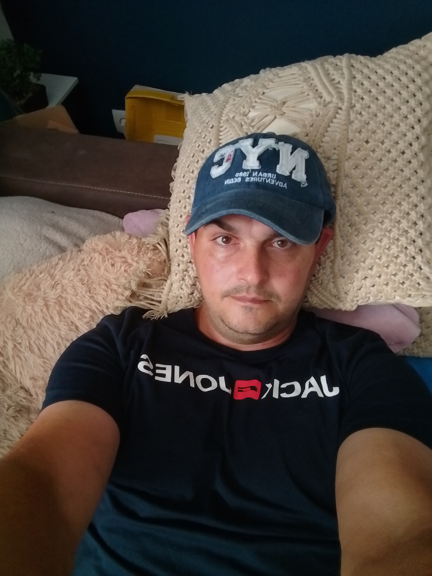 A man wearing a blue baseball cap and a black t-shirt with "JACK & JONES" printed on it is lying on a sofa. The cap has "NYC" printed in white. He is resting his head on a beige crocheted pillow. To his left, a fluffy, cream-colored stuffed animal is visible. In the background, a dark blue wall and a white shelf with a small green plant are seen. The overall environment suggests a casual indoor setting. There is no information available about the location in Wedel, Germany.