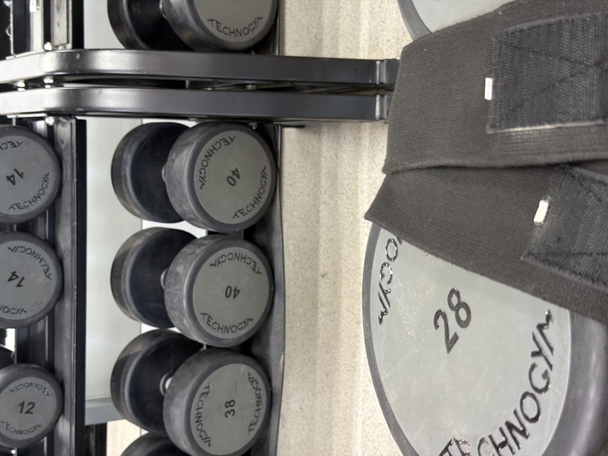 The image depicts a gym setting in Chaikovsky, Russia, specifically focusing on a rack of dumbbells and a weight plate. In the foreground, a large, gray, circular weight plate with the branding "TECHNOGYM" and the number "28" embossed on it is visible. A black fabric strap, likely a wrist or ankle wrap, with Velcro fasteners is partially obscuring the weight plate. In the middle ground, a tiered rack holds several dumbbells. The top shelf displays dumbbells with weights marked "12" and "14". The shelf below it contains dumbbells with weights marked "38" and "40". The dumbbells are predominantly black with gray circular ends where the weight is indicated. The background shows the metallic structure of the dumbbell rack and the gym floor, which appears to be a light-colored, textured surface. The lighting suggests an indoor environment with overhead illumination.