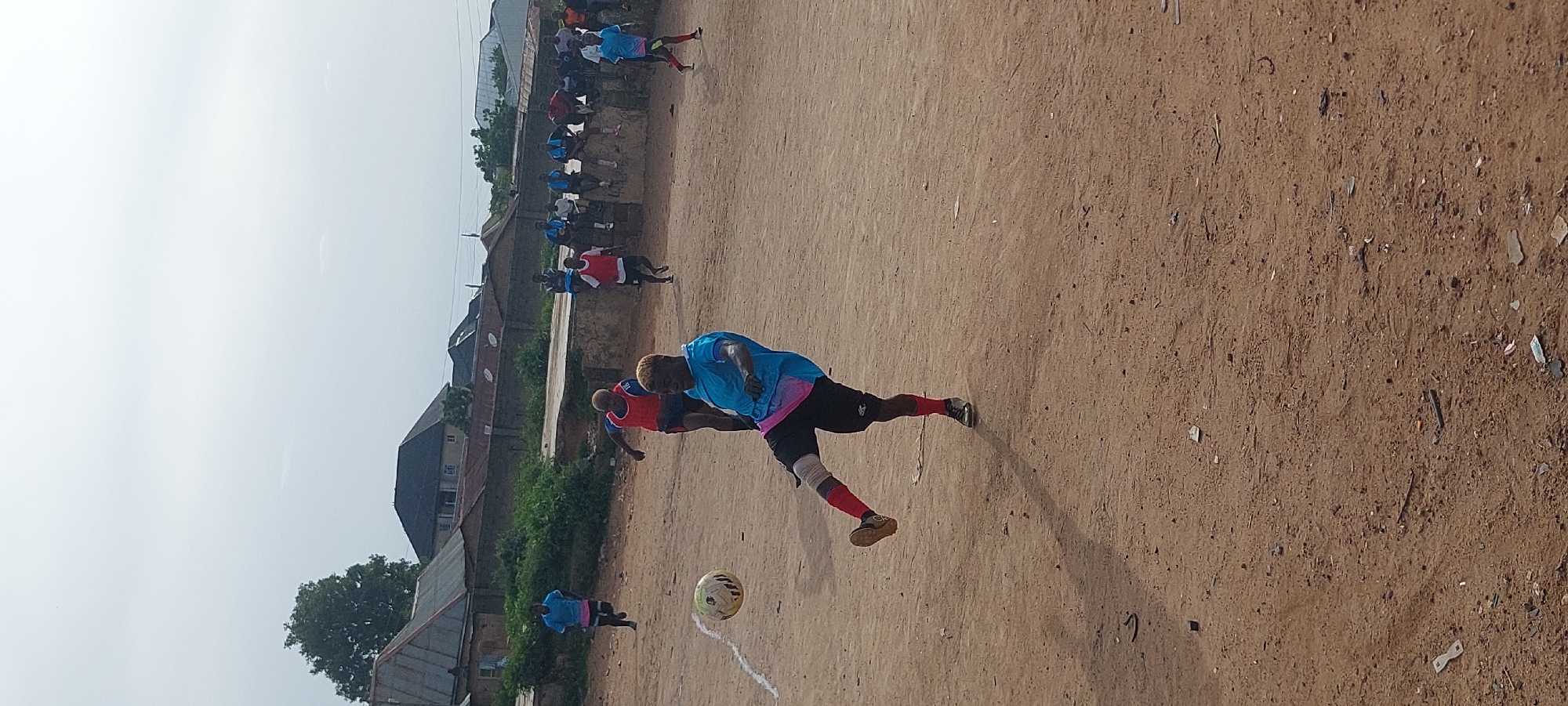 A full shot shows a soccer game in progress on a dirt field in Bukuru, Nigeria. In the foreground, a male soccer player with bleached blonde hair, wearing a blue and pink jersey, black shorts, and red socks, kicks a white and yellow soccer ball. The player is positioned in mid-air, with his right leg extended to connect with the ball. In the middle ground, other players in blue and red jerseys are visible on the field. In the background, a group of spectators sits on a low concrete wall. Residential buildings with corrugated metal roofs line the distant horizon, under a hazy sky. The dirt field is uneven and littered with debris.