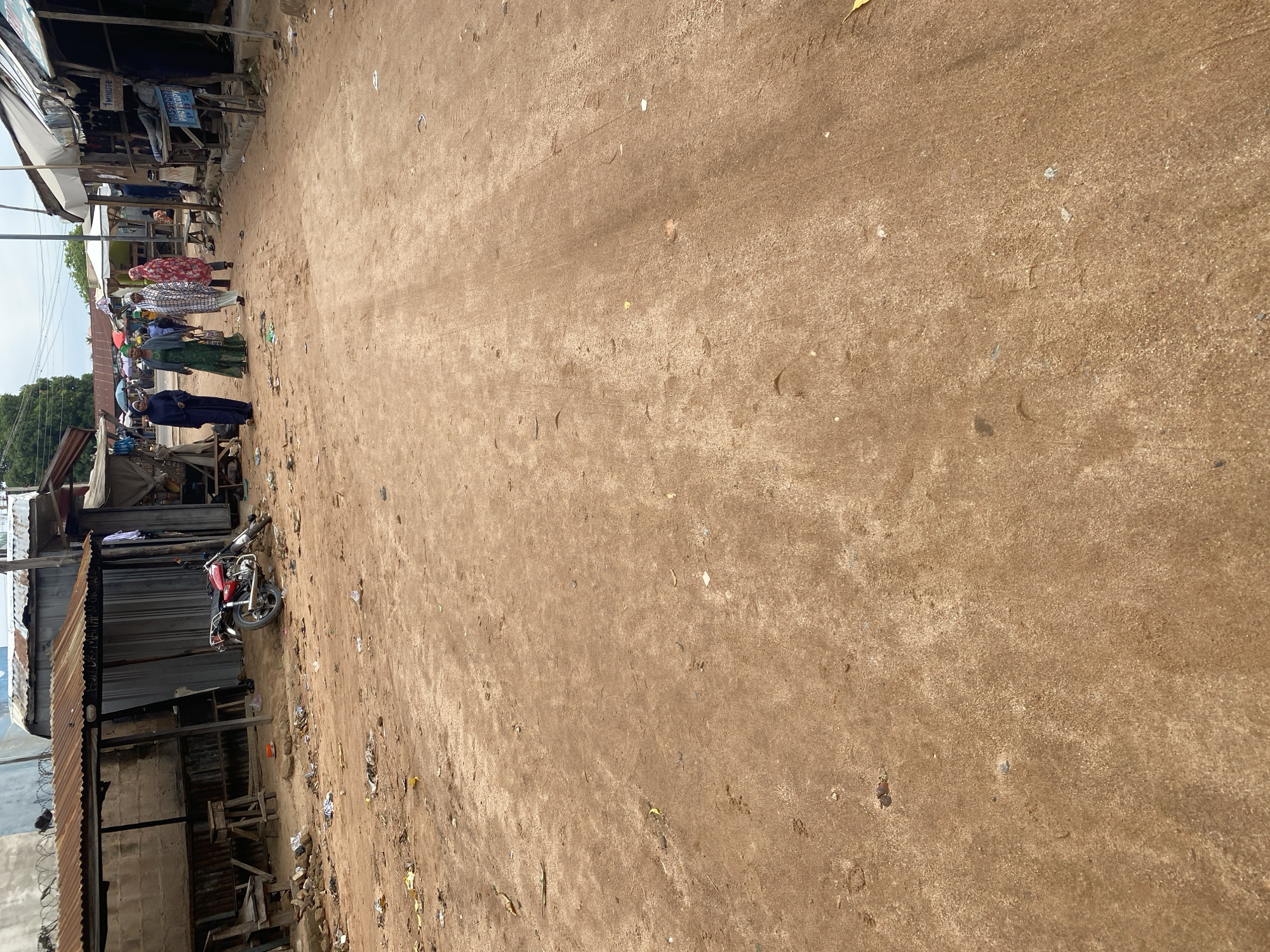 This media file depicts a street scene in Jalingo, Nigeria. The image is oriented vertically, showing a dusty, dirt road with various structures and people along its edge.
The subjects include several individuals, some of whom are wearing traditional clothing like hijabs and flowing garments, suggesting a cultural context. They appear to be standing or walking along the side of the road. Objects present include a red motorcycle parked near a corrugated metal structure, and makeshift stalls or shops constructed from wood and metal. Debris and litter are scattered across the road and the surrounding area, indicating a less-than-pristine environment. The buildings appear to be simple, functional structures, possibly shops or residences.
The activity appears to be ordinary street life, with people going about their day. There's no indication of a specific event taking place; it seems to be a candid snapshot of daily existence.
Notable details include the dusty, dry terrain, typical of a sunny climate. The lighting suggests it could be daytime, likely late morning or afternoon given the shadows. The presence of the motorcycle and the types of structures hint at a developing or less urbanized area.
There is visible text on a sign in the background, partially obscured, which appears to read "1 MINUTE" and another section with "ASTROLOGY BIRTHDAY POS". This suggests some form of commercial activity or service being offered.