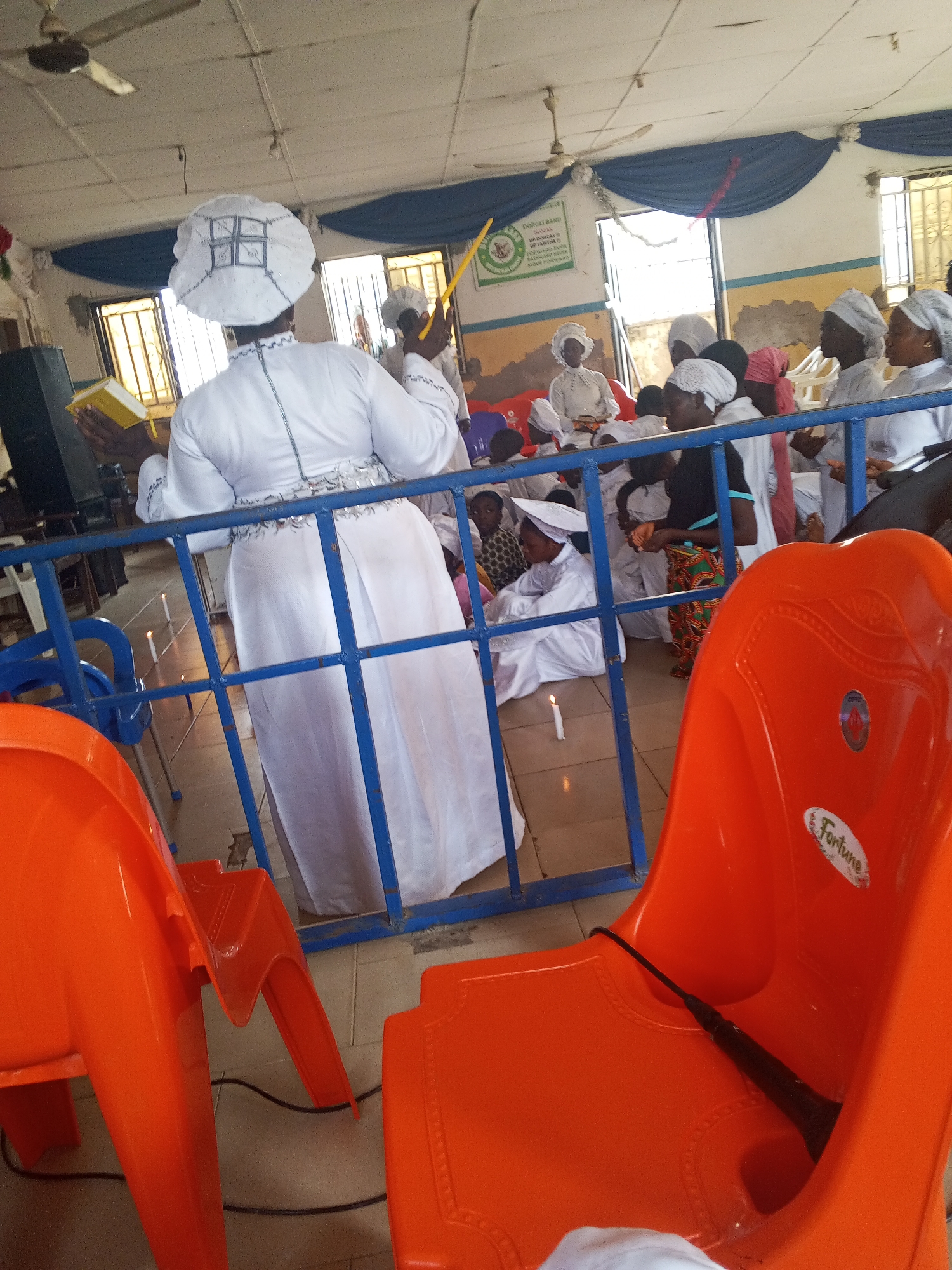 The image captures a gathering of people inside what appears to be a church or religious setting in Lokoja, Nigeria. Most individuals are wearing white clothing, with some specifically dressed in white robes and head coverings, suggesting a religious event or service. A woman in the foreground stands behind a blue metal railing, holding a yellow book and a long yellow object, possibly a candle. Several candles are placed on the floor near the railing. Other people are seated or kneeling in the background. Orange plastic chairs are visible in the foreground, with one prominently displaying the word "Fortune." The location has ceiling fans, and there is a banner hanging in the background. The building's interior shows some signs of wear.