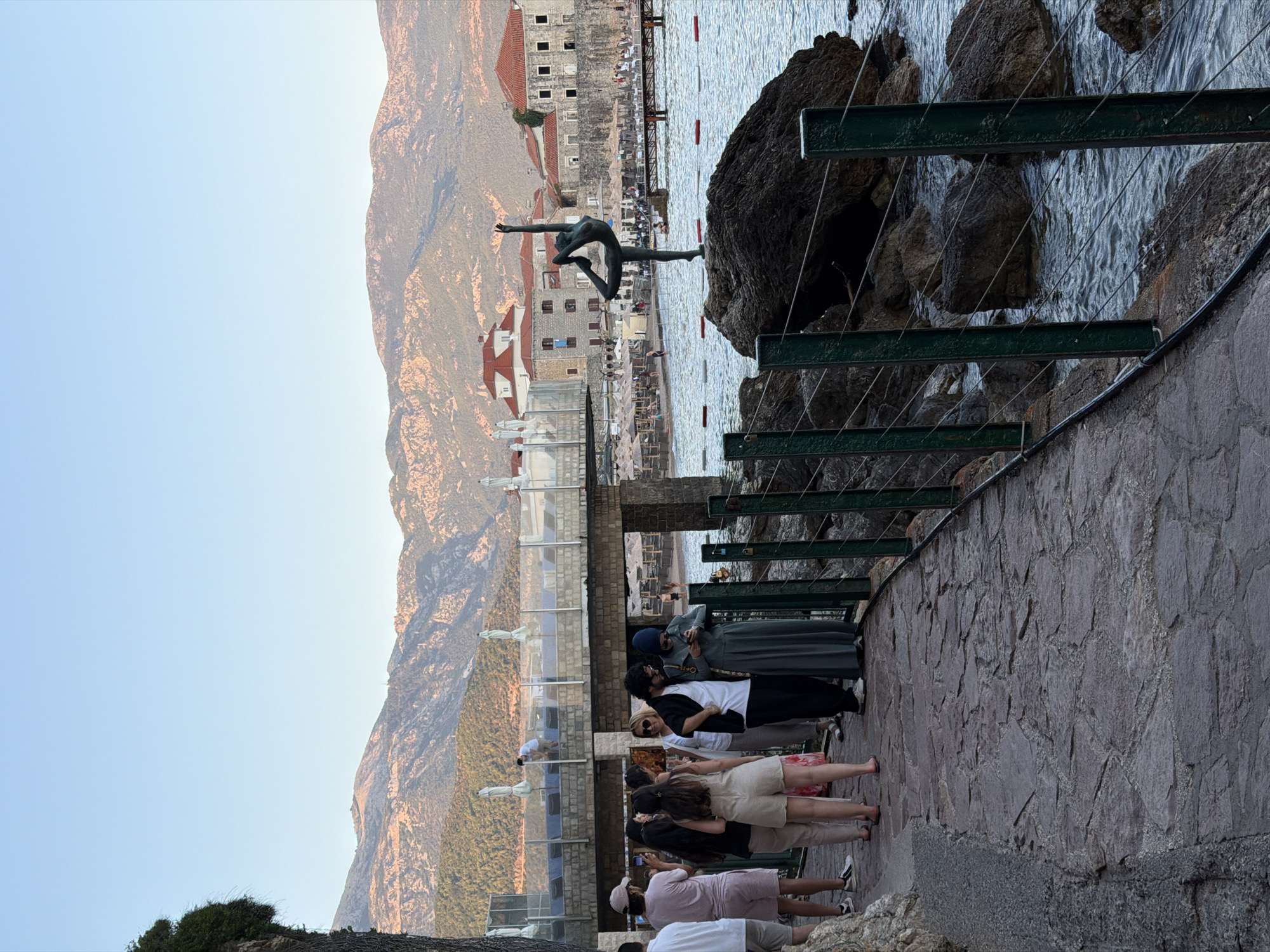 The image depicts a scenic promenade in Budva, Montenegro, with the Adriatic Sea on the right and a mountainous backdrop. In the foreground, a stone pathway runs alongside a green metal fence with wires. Several people are walking and standing on the pathway. To the right, a dark rock in the sea supports a bronze statue of a dancer in a dynamic pose. The sea has gentle waves, and red and white buoys mark an area. Further back, a beach with umbrellas and lounge chairs is visible, along with buildings with red tile roofs characteristic of Budva's Old Town. A bridge spans over a section of the water, and a few people can be seen on it and on the beach. The sky is clear and light blue, suggesting it is either early morning or late afternoon. The lighting on the mountains indicates a warm, golden hour.