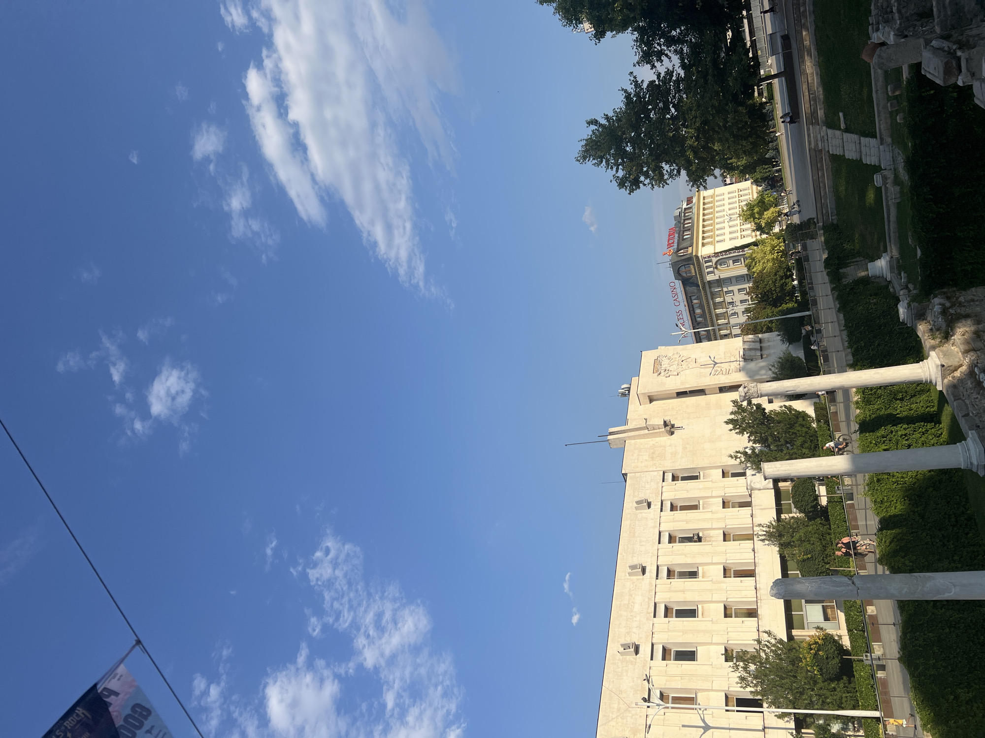 The image depicts an outdoor scene in Plovdiv, Bulgaria, under a clear blue sky with scattered white clouds, suggesting it's a sunny day, likely daytime. The perspective is from a slightly elevated viewpoint, looking towards a modern building with a light-colored facade and multiple windows. In front of this building, there are remnants of ancient structures, including large stone columns and weathered stones, hinting at a historical site. Greenery, including trees and manicured bushes, surrounds the ancient ruins and lines the pathway in front of the modern building.
In the mid-ground, people can be seen walking along a path, some individuals are sitting on benches, and a person is riding a bicycle. The modern building to the right displays text at the top, reading "VICTORIA" in large red letters, and below it, "PRINCESS CASINO" in smaller red lettering. Further in the background, another building with a pale yellow facade and numerous windows is visible, with more trees interspersed. A banner is partially visible in the lower left corner, with some text that appears to be "000" and "ROCK". The overall impression is a blend of historical significance and modern urban life.