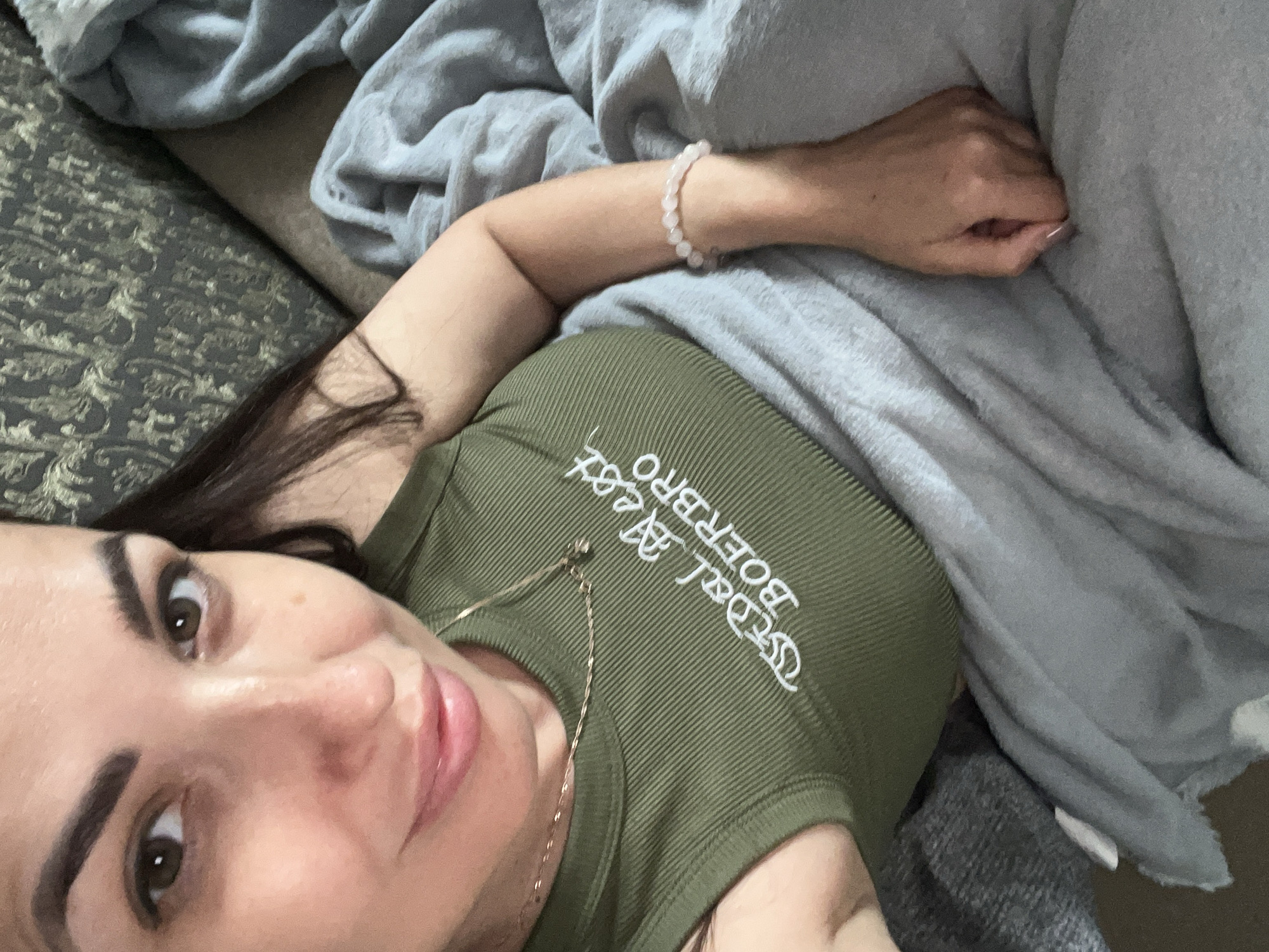 A woman is lying down on a sofa, covered by a light blue blanket. She is wearing a green tank top with white lettering that reads "Dal Neer Boerbro". A gold necklace is visible around her neck, and she is wearing a light pink beaded bracelet on her right wrist. The background shows a patterned sofa and more blankets. The lighting is natural, and the image appears to be a selfie.