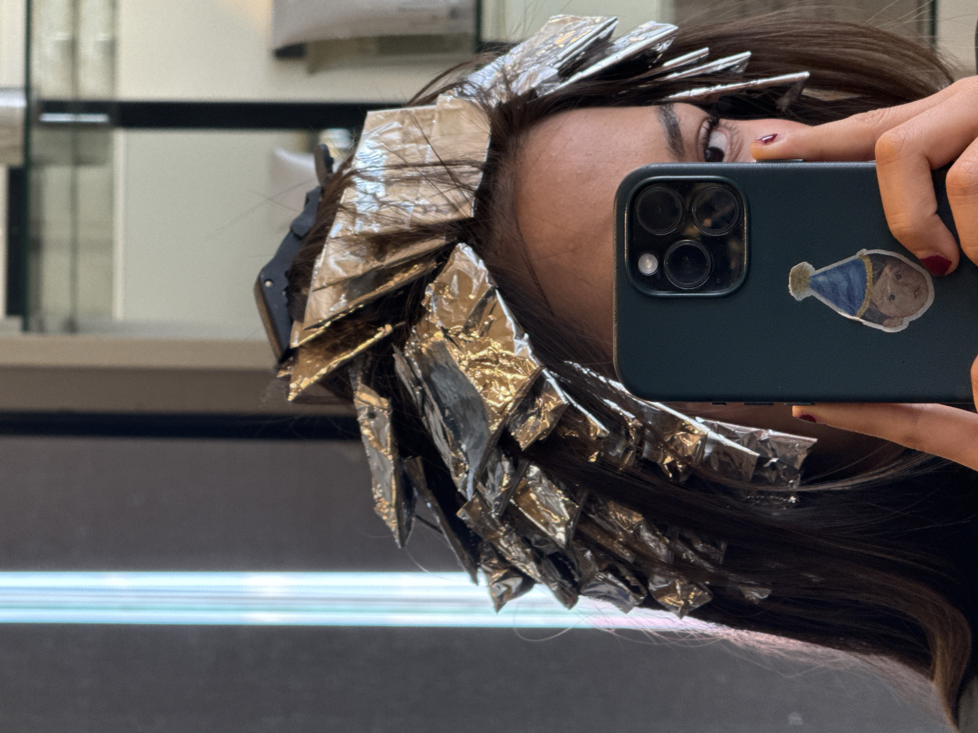 The image shows a person with dark brown hair having highlights applied, as evidenced by the foil in their hair. They are holding a dark-colored smartphone with three camera lenses and a sticker of a bear wearing a party hat. The person is partially visible in the reflection of a mirror. The background shows a blurred interior, possibly a hair salon or similar establishment. The location details of Unknown, Unknown are provided, but without further context, it's impossible to confirm the image's location.