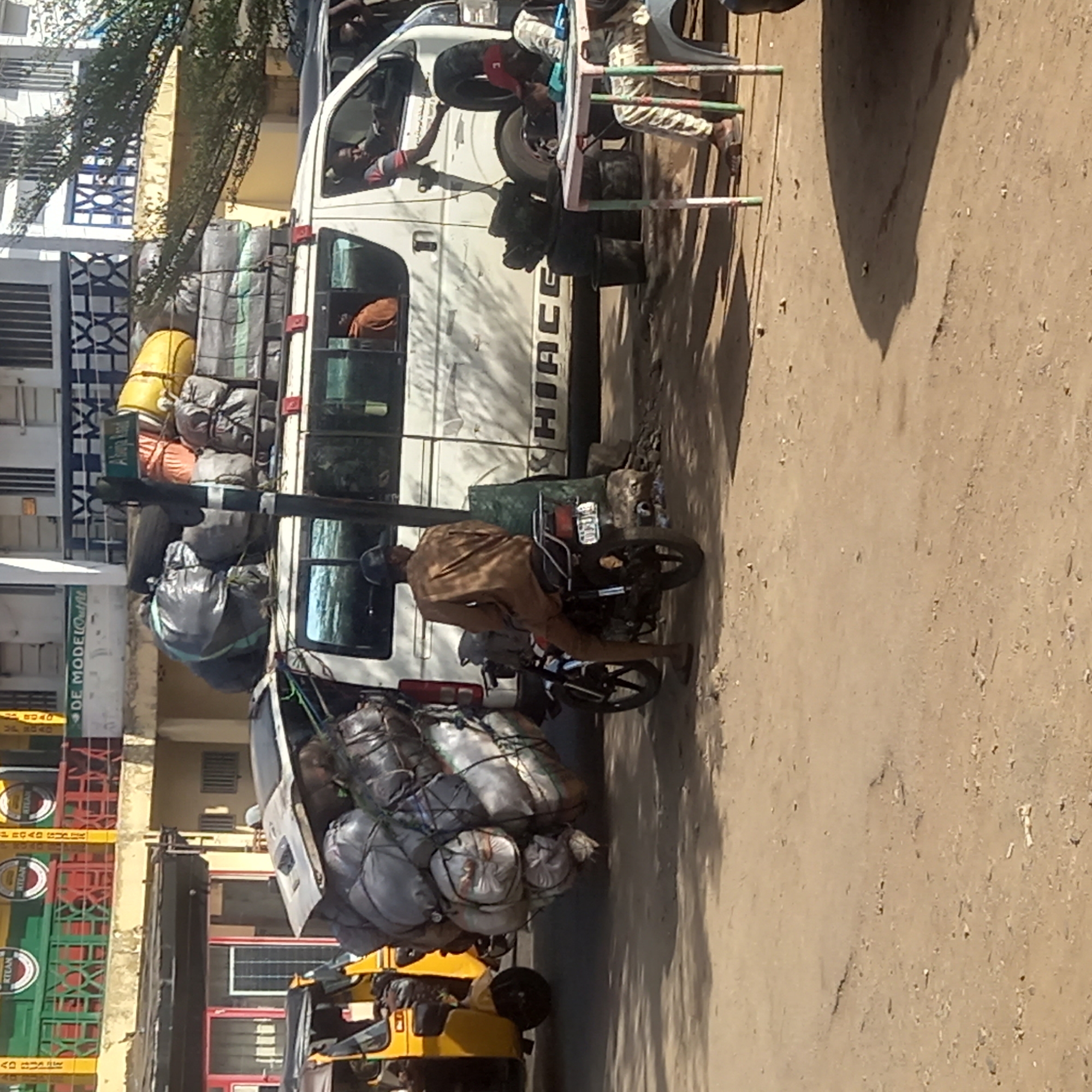 The scene is a street in Dadi Riba, Nigeria. A white Toyota HiAce van is parked on the side of the road, heavily loaded with luggage on the roof. A man on a motorcycle wearing a brown outfit is stopped next to the van. A yellow tricycle taxi is parked further down the street, also heavily loaded with luggage. Pedestrians are seen around the van, interacting with it. Buildings line the street, including one with a sign that reads "DE MODE@Outfit." The ground is a dirt and paved surface.