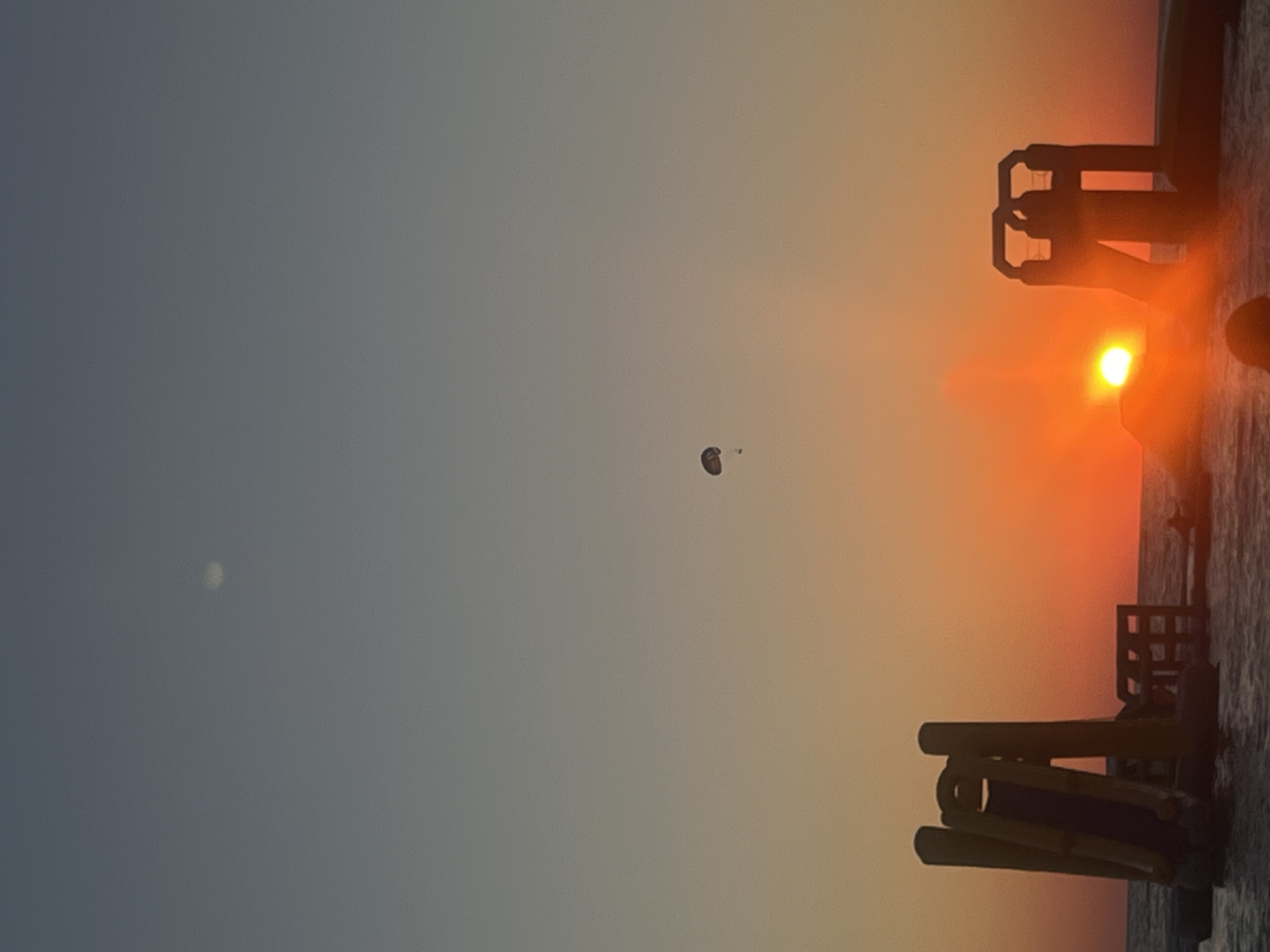 A parasailer is suspended in the air above the ocean during sunset in Sochi, Russia. The sky transitions from a dusky blue at the top to a vibrant orange near the horizon, where the sun is setting. In the foreground, the silhouettes of yellow inflatable water slides are visible on the water. A faint circular object, possibly the moon, is visible in the upper portion of the sky.
