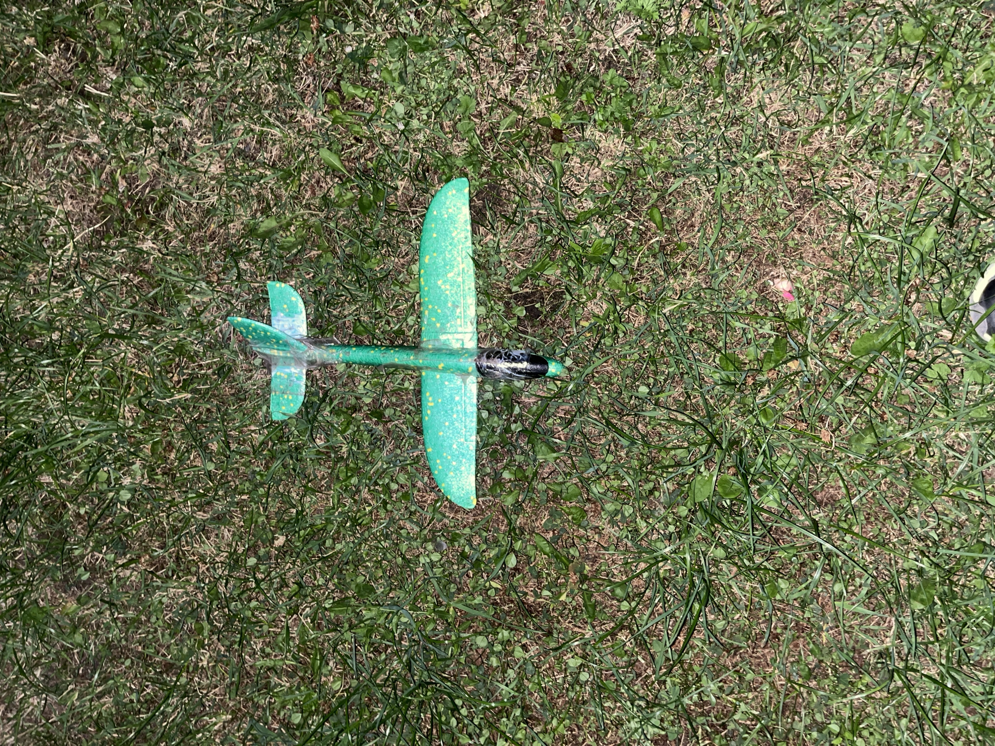 An overhead, medium shot shows a green toy airplane with yellow speckles lying on grass. The airplane is positioned vertically in the center of the frame, with its nose pointing down. The grass is a mix of green and dry blades, with some patches of dirt visible. The lighting suggests it is daytime. There is no indication of the location within the city of Saratov, Russia, or any people, actions, or interactions.