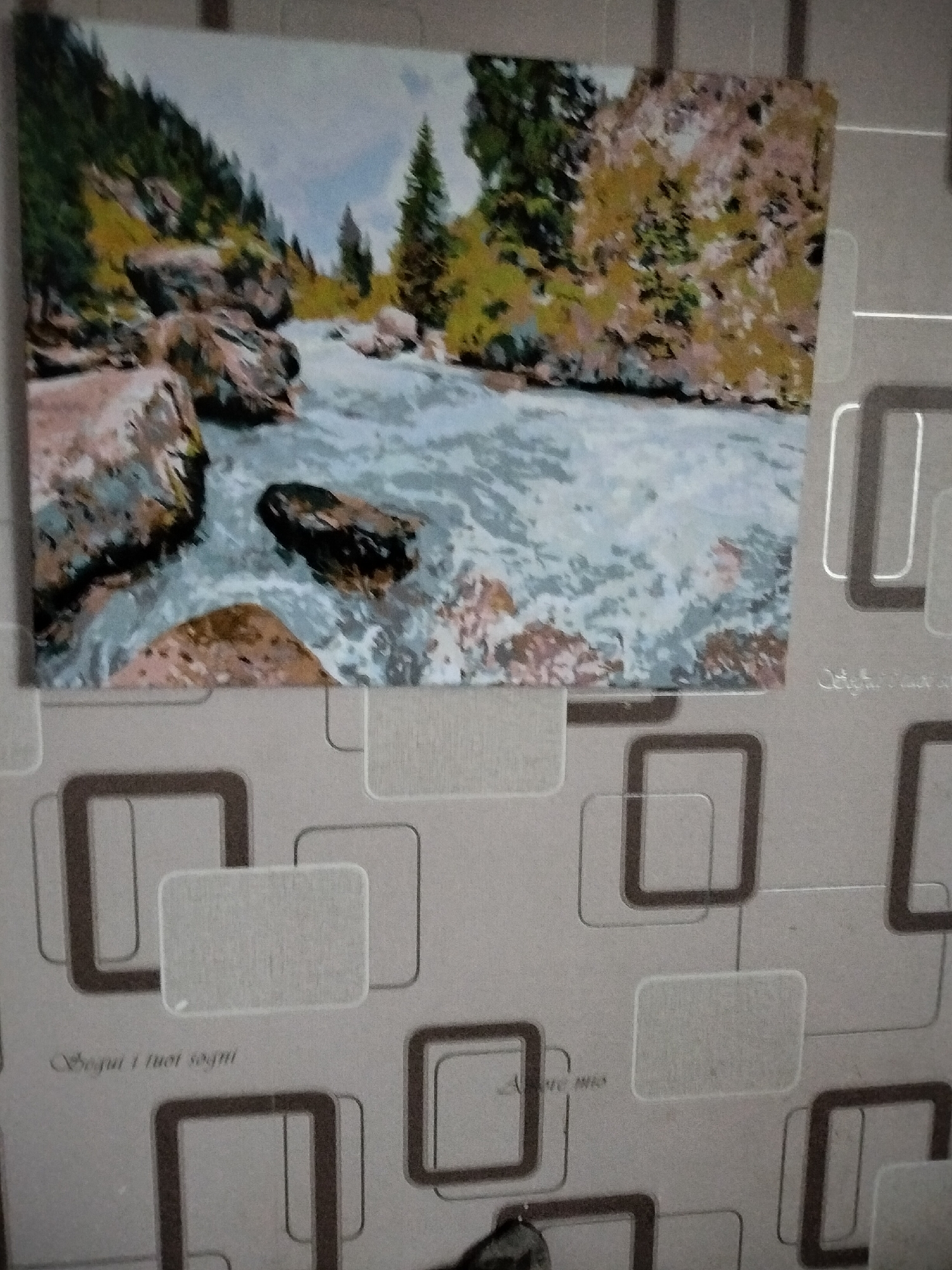 The image shows a painting of a river flowing through a rocky, mountainous landscape. The painting is hanging on a wall with a patterned wallpaper. The wallpaper features a repeating design of interconnected squares and rectangles in shades of gray and brown. There are also some areas of lighter gray that appear to be textured. Below the painting, on the wallpaper, there are two instances of text written in a cursive font. The first reads "Segui i tuoi sogni" and the second reads "Amore mio." The image does not contain any information that would allow for identification of its location in the city of Nizhnedonskoy, Russia, or any specific details about people, objects, or actions.