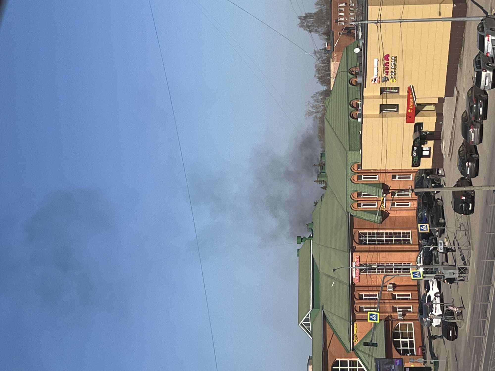 The image shows a street scene in Klin, Russia. Buildings with green roofs and brick facades line the street. A yellow building with various signs is also visible. Several cars are parked along the side of the street. In the distance, smoke is rising into the blue sky. Overhead wires crisscross the sky.