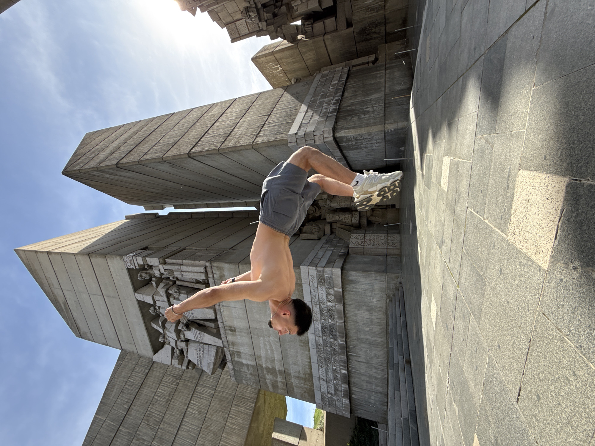 The image shows a man performing a backflip in front of the Monument to 1300 Years of Bulgaria in Shumen. The monument is composed of large, stylized concrete blocks with inscriptions and reliefs. The man is wearing gray shorts and white sneakers. He has his arms extended. The day is sunny.