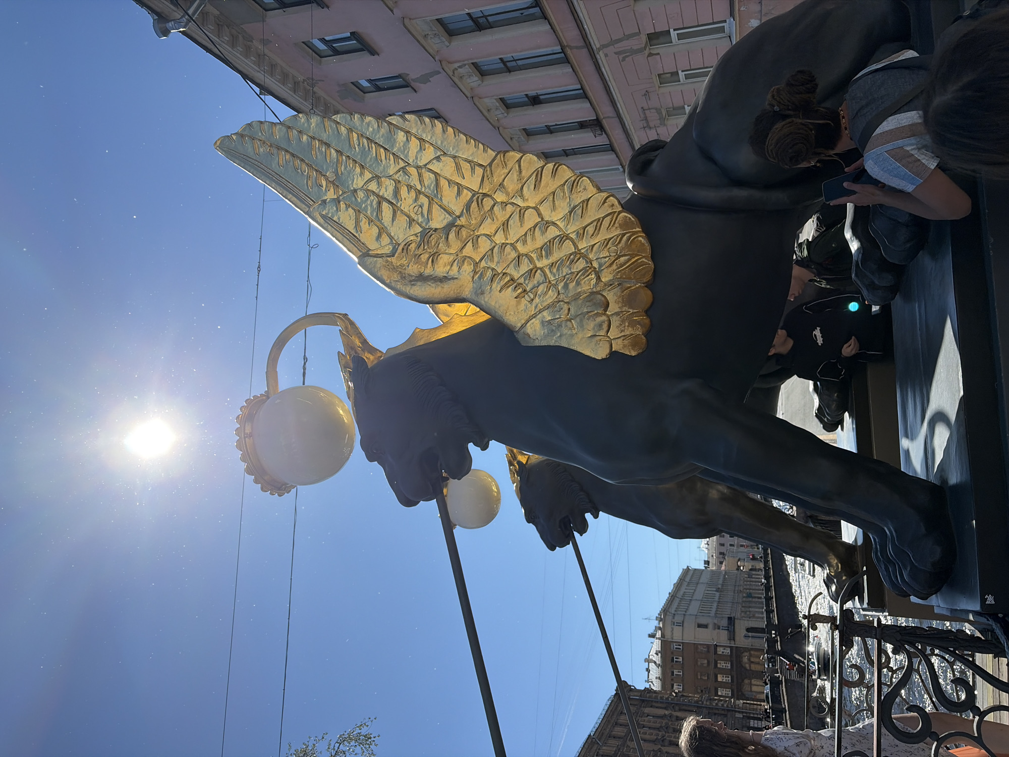 The image depicts the Griffins of Bank Bridge in Saint Petersburg, Russia, on a bright, sunny day. The Griffins, sculpted from bronze and with gilded wings, are situated on a bridge that spans a canal. The sun is high in the bright blue sky, casting a glare and creating lens flare. In the foreground, the statues of the Griffins dominate the scene, their large bronze bodies and impressive gilded wings capturing the viewer's attention. Two lamps with white globes are mounted on ornate posts above the Griffins. Several people are visible in the scene. On the left, a woman with long curly hair, wearing a white dress, stands near a decorative railing, looking towards the canal. In the middle ground and background, people are gathered around the statues. On the right side, a woman with her hair in a bun is holding a smartphone, appearing to take a photo or video. In the canal below, a boat is navigating the waterway. The buildings lining the canal are typical of Saint Petersburg's architectural style, with multi-story structures featuring windows and decorative elements. The overall atmosphere is one of a lively tourist destination on a clear, warm day.