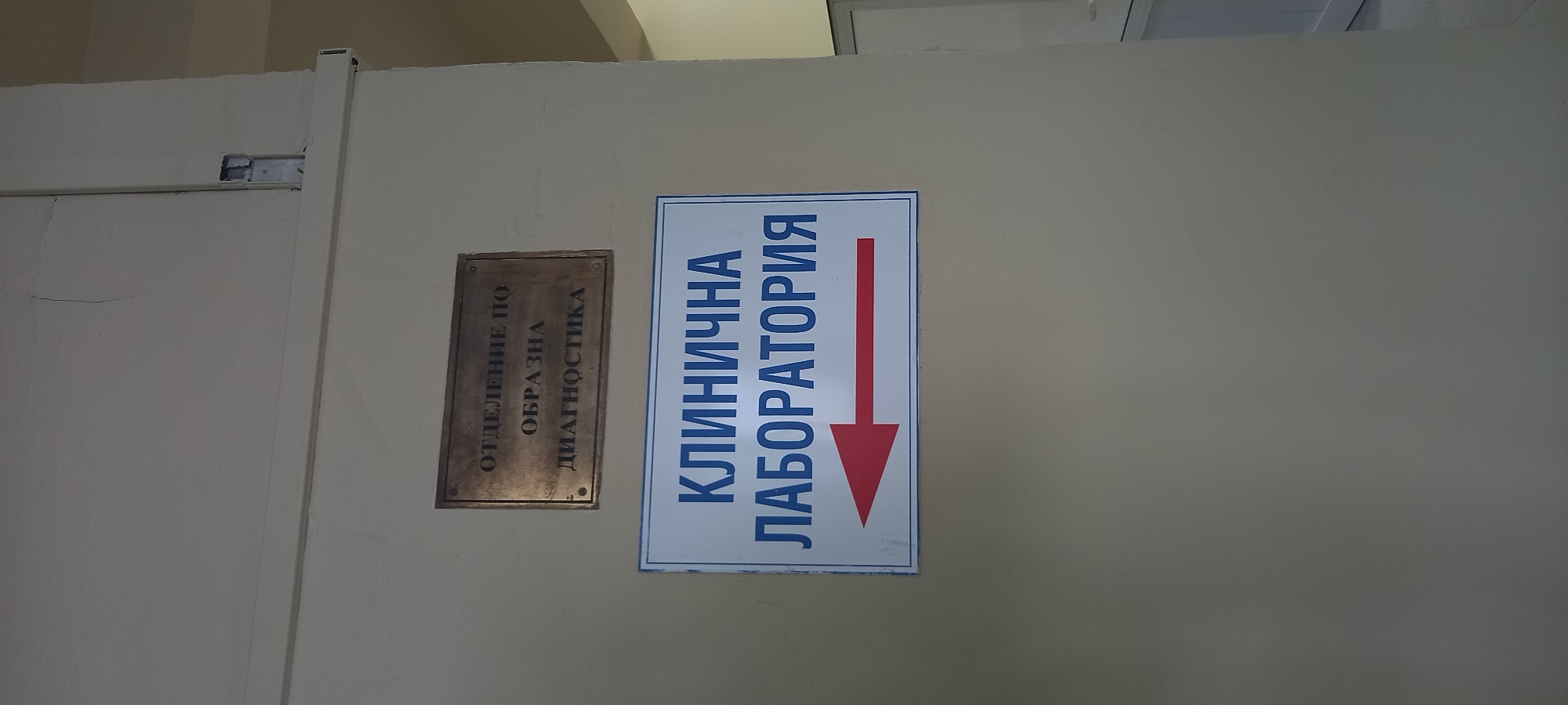 The image shows a wall in an indoor setting, likely a hospital or clinic. There are two signs on the wall. The top sign is a dark, rectangular plaque with text in Russian. The text reads "ОТДЕЛЕНИЕ ПО ОБРАЗНА ДИАГНОСТИКА," which translates to "Department of Imaging Diagnostics." Below this, there is a white rectangular sign with a blue border. This sign also has text in Russian, "КЛИНИЧНА ЛАБОРАТОРИЯ," meaning "Clinical Laboratory," and a red arrow pointing to the left. The wall appears to be painted a light, neutral color. The environment seems to be a hallway or corridor, as suggested by the presence of doors or openings in the background on the right side of the image. There are no people visible in the image. The image does not provide any information or context to suggest a location in Silistra, Bulgaria.