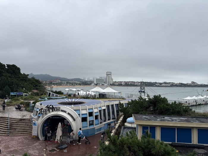 This image captures a coastal scene on an overcast day, specifically depicting the Qingdao Seal Museum in Shinan District, Qingdao, China.
In the foreground, the "SEAL MUSEUM" (海豹馆) is a dominant structure, semi-circular in design with a light blue and white exterior. Its roof is extensively covered with radially arranged solar panels. A large arched entrance is visible on the left side of the building, where approximately six individuals are observed entering or exiting. One person is pushing a stroller. A dark paved plaza extends in front of the museum, and to its left, a multi-tiered stone stairway leads up to a higher vantage point where a few people are seated or standing. To the lower right, part of a rectangular building with a dark roof and blue-paneled exterior is visible, partially obscured by green coniferous trees.
The midground features a large body of water, likely Fushan Bay, with a lengthy pier or floating walkway extending from the right, supporting multiple white, tent-covered structures. A prominent metal framework, possibly a diving platform or suspended structure, is positioned over the water. Along the distant shoreline, a sandy beach is visible, populated with numerous white beach umbrellas and tents, indicating a recreational area.
The background reveals a dense urban landscape characterized by numerous buildings, including a cluster of high-rise structures, one tall white skyscraper with a distinctive finial. Beyond the city, rolling green hills and mountains are visible under a uniformly gray, overcast sky, indicating cloudy weather conditions.