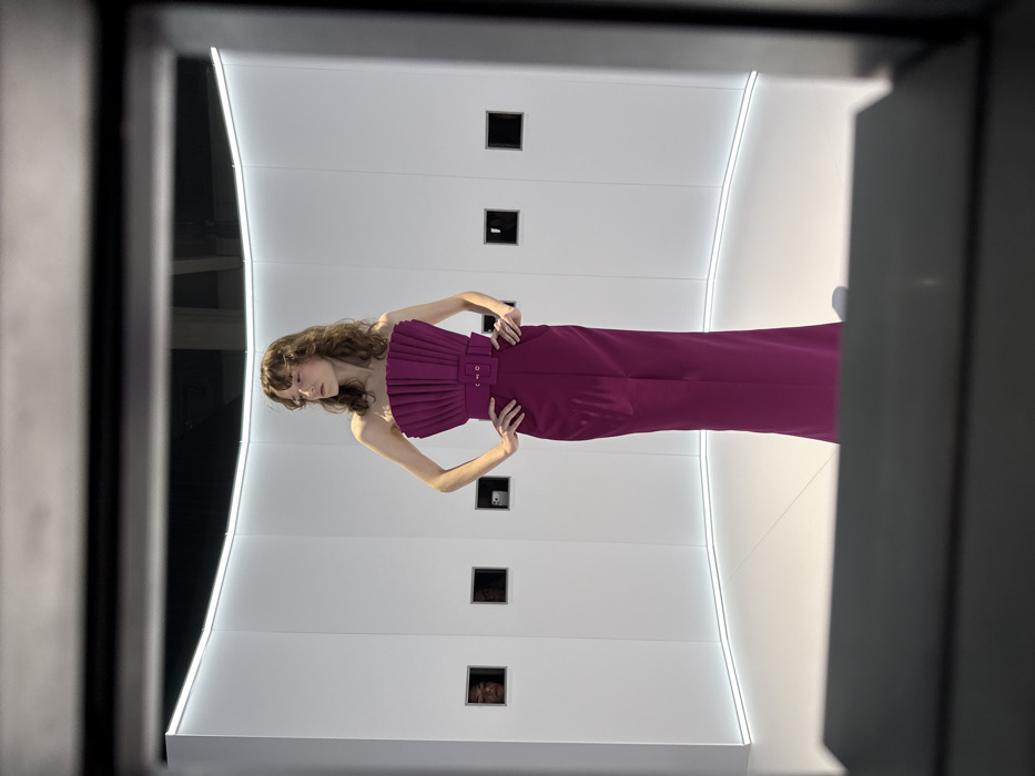 A woman in a magenta strapless jumpsuit poses with hands on hips against a stark white backdrop. The jumpsuit features a ruffled bodice and a belted waist. She has brown curly hair. The backdrop is vertically paneled with four small square cutouts, each seemingly containing a small object. The overall scene is framed by dark gray vertical structures, suggesting a doorway or enclosure. A curved light fixture runs along the edge of the white backdrop.