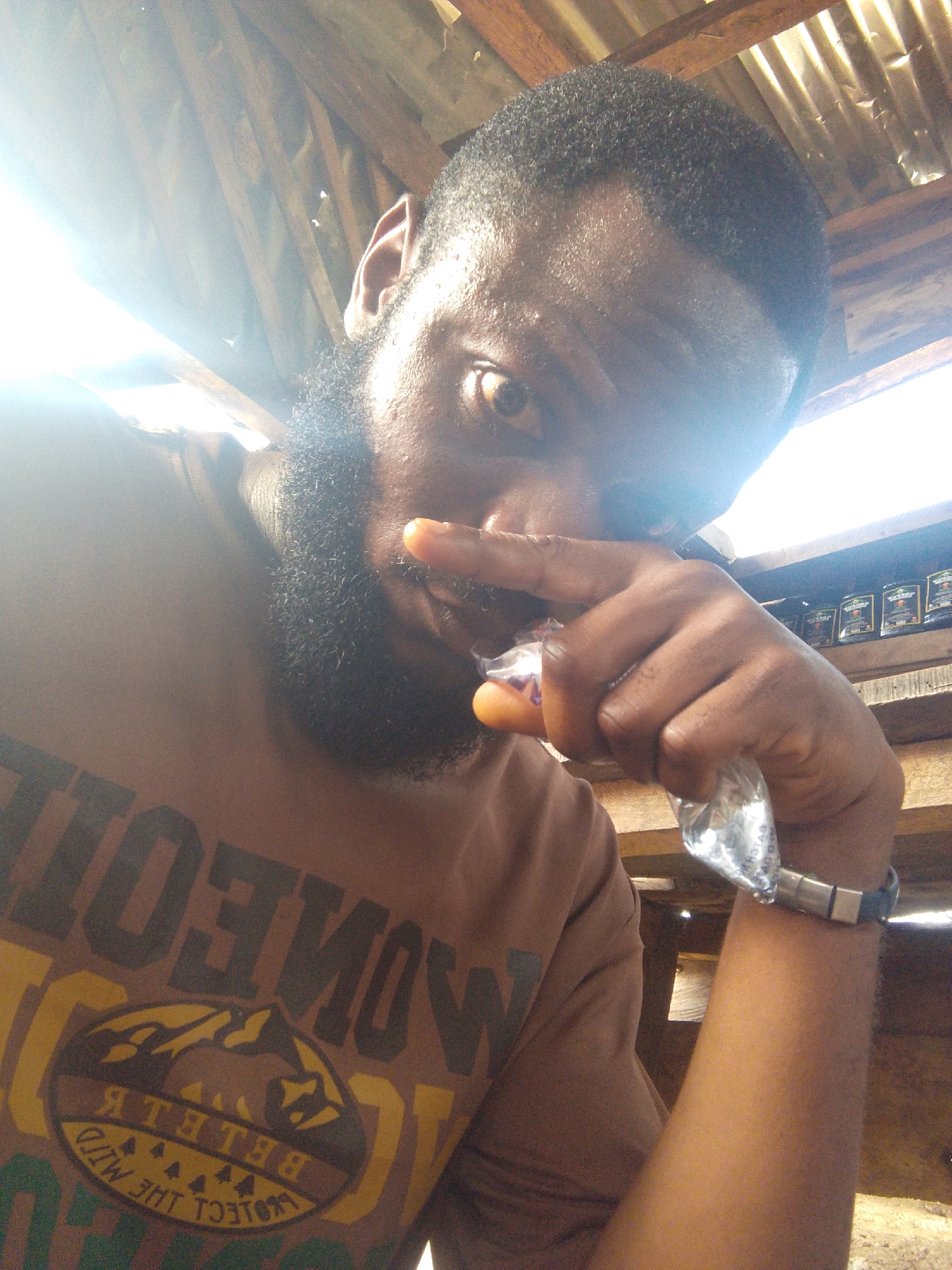 A man with dark skin, a beard, and short hair is holding a small, clear plastic bag near his mouth. He is wearing a brown t-shirt with a graphic design that includes the words "MONEO" and "PROTECT THE WILD." A bracelet is visible on his wrist. The setting appears to be an indoor space with a corrugated metal roof. A shelf with packaged goods is in the background. The location is unspecified within Lokoja, Nigeria.