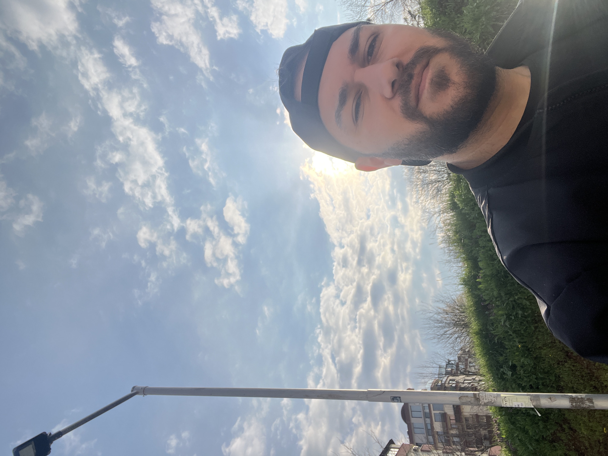 A selfie taken in Veliko Tarnovo, Bulgaria, shows a man with a beard and a black cap. The sky behind him is partly cloudy, with the sun shining brightly. Below the sky, there is a streetlight pole. In the background, one can see some bushes, trees, and the upper part of a multi-story building.