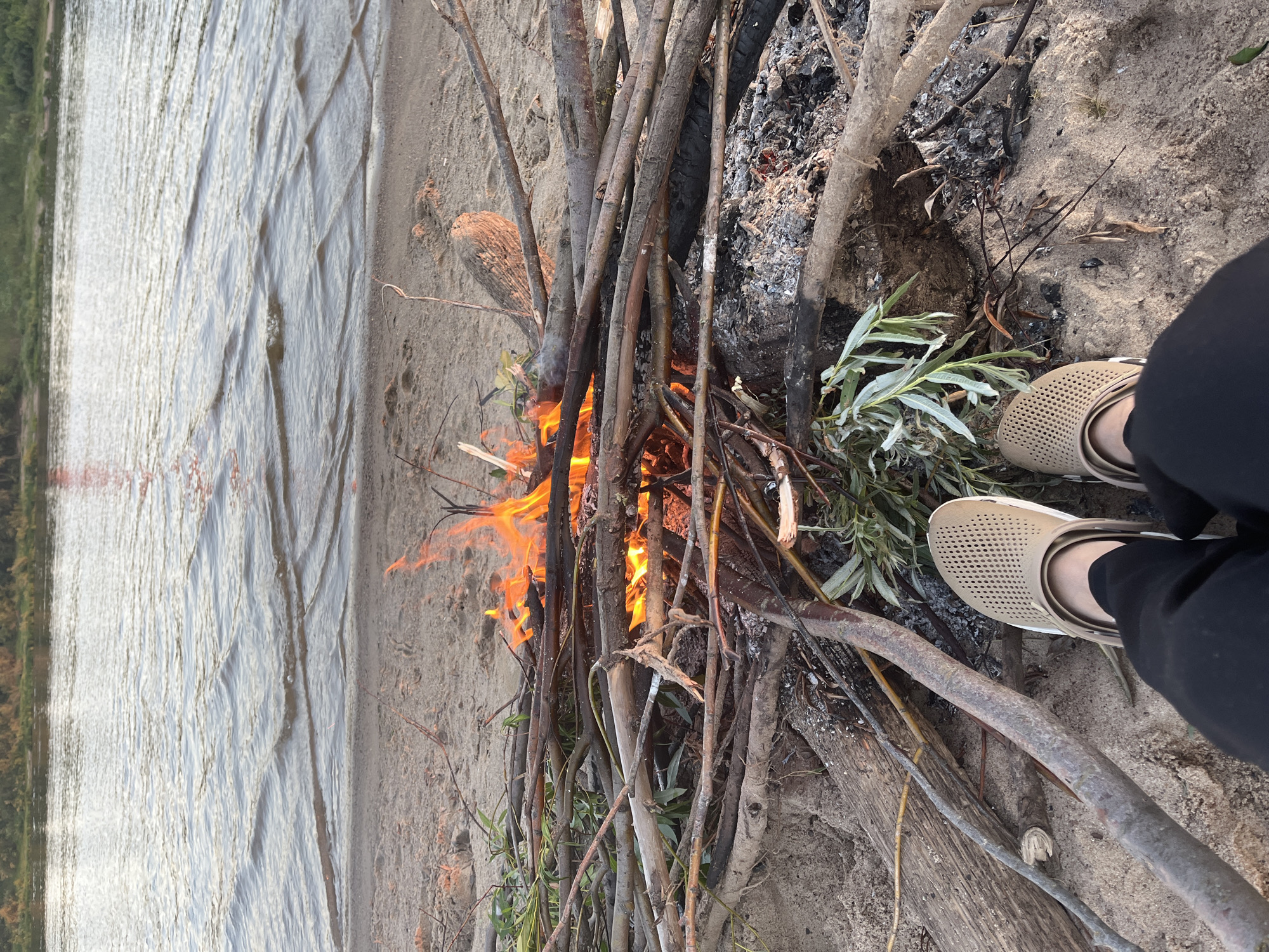 The image shows a view from ground level looking towards a river with a bonfire on the sandy bank in the foreground. Two feet wearing light brown perforated clogs and dark pants are visible at the bottom of the frame, facing away from the viewer. The bonfire is actively burning, with orange flames and smoke rising from a pile of dry branches and logs. The river is calm with small ripples, and the far bank is lined with green vegetation. The overall scene suggests a peaceful outdoor setting, possibly for a picnic or camping trip. The specific location in Nizhny Novgorod, Russia, cannot be determined from the visual information alone.