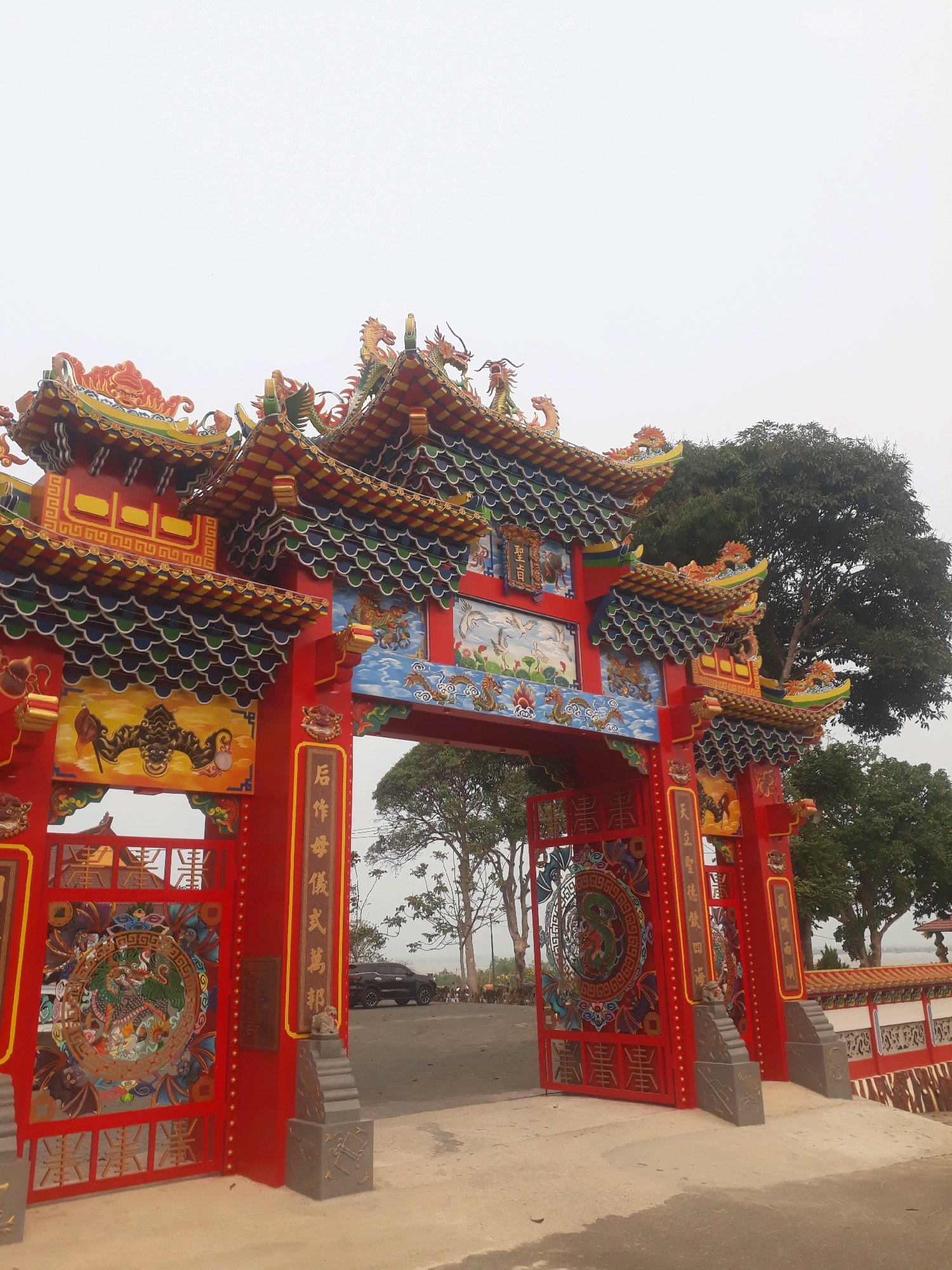 This image depicts the entrance to a vibrant Chinese temple, likely located in Pangkalpinang, Indonesia, judging by the architectural style and common presence of such structures in areas with significant Chinese-Indonesian communities. The entrance is a grand, multi-tiered structure painted in bright red, adorned with intricate dragon motifs, traditional roof tiles in green and yellow, and detailed paintings on panels.
The central archway features a red gate with ornamental carvings, including a prominent circular motif with a dragon on the left gate. The gateposts are decorated with calligraphy and small guardian lion statues at their bases. Above the archway, a painted mural depicts mythological scenes, possibly with cranes and dragons, set against a blue sky. The roofs are elaborately decorated with colorful, curved tiles and more dragon sculptures positioned along the ridges and corners, symbolizing power and good fortune.
To the right of the entrance, a stone wall with a tiled roof borders the temple grounds, and behind it, lush green trees provide a natural backdrop. A dark-colored SUV is parked on a paved area visible through the open gate. The sky is overcast, suggesting either an early morning or a hazy day. The ground in front of the entrance is a light-colored, paved surface. The overall impression is one of a richly decorated and culturally significant religious site.