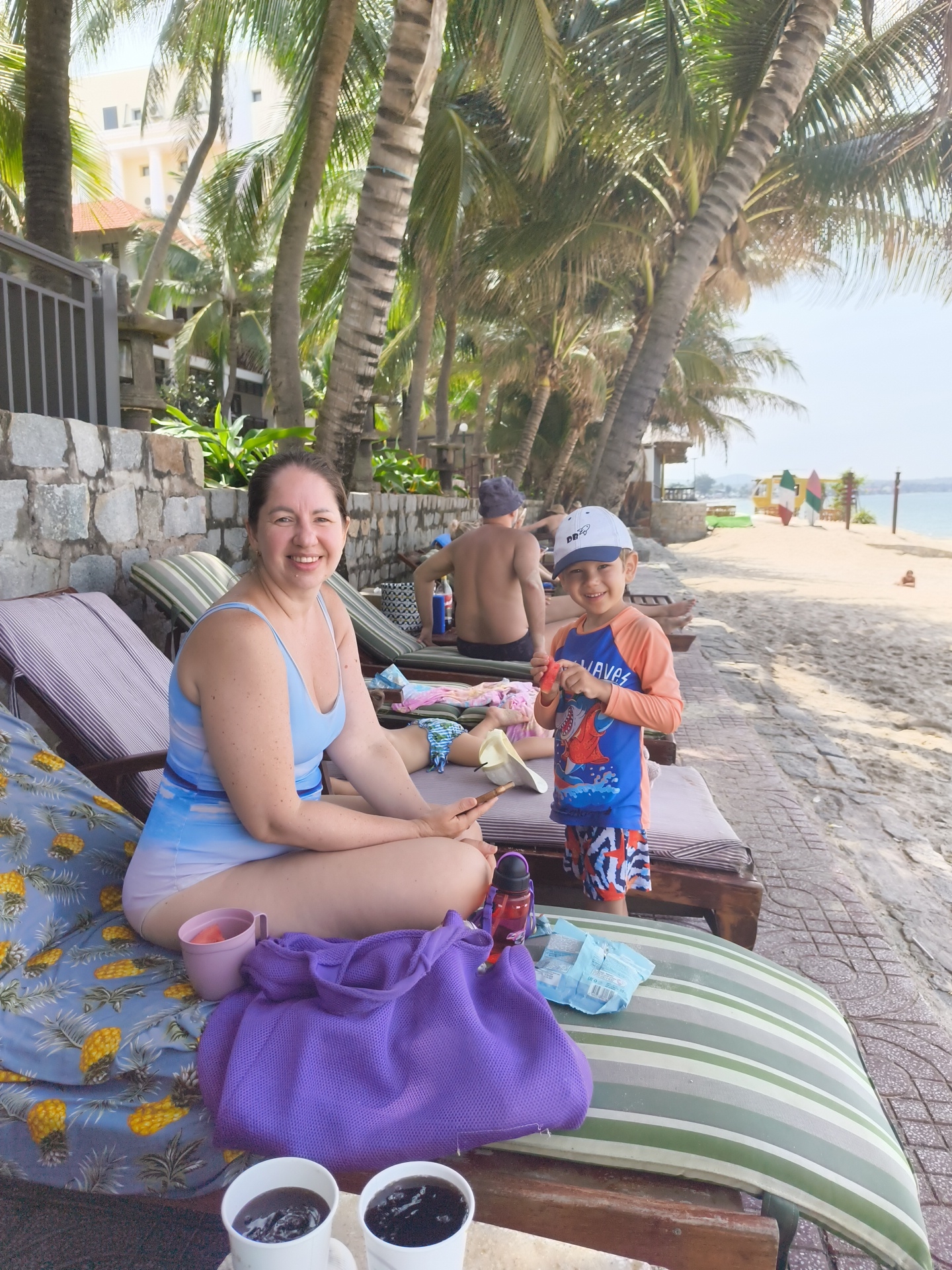 In Phu Thuy Ward, Vietnam, a woman in a blue and white swimsuit sits on a striped lounge chair on a beach. She is smiling and holding a phone. Next to her is a young boy wearing a blue and orange rashguard and patterned shorts, holding a piece of fruit and smiling. In front of them, on the lounge chair, are a purple bag, a purple cup, and two white cups with dark liquid. In the background, palm trees line the sandy beach, with a resort building visible behind them. Another person is seen sitting on a lounge chair further down the beach.