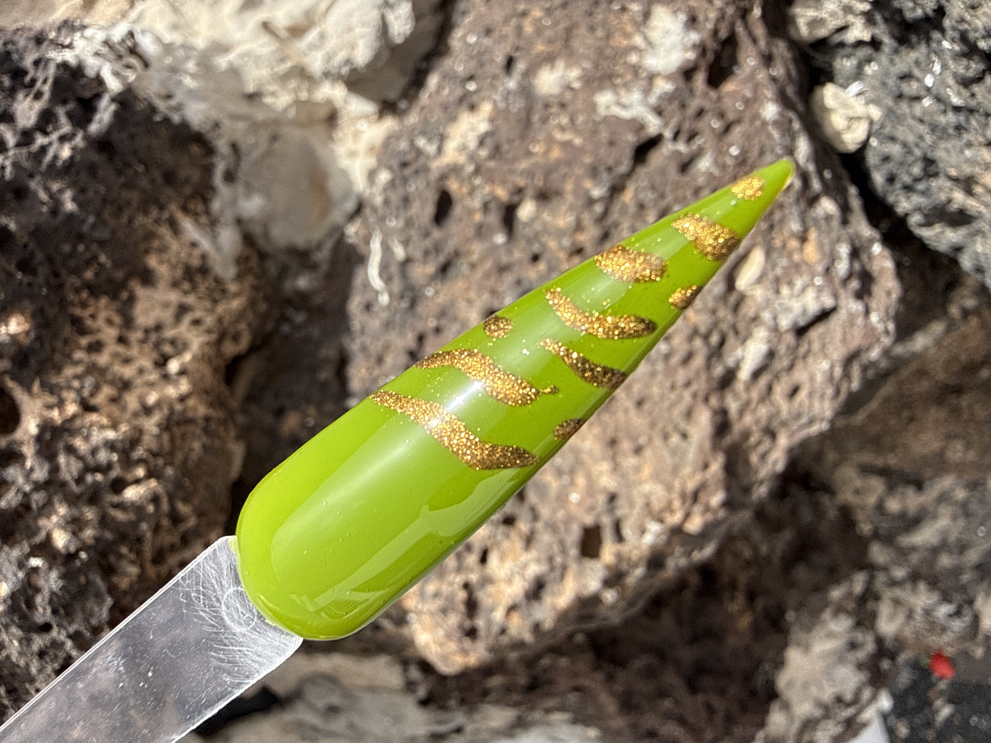 A close-up shows a lime green nail painted with horizontal lines of gold glitter. The nail is held on a clear plastic stick. The background appears to be a textured rock surface, possibly volcanic rock, which is common in Yaiza, Spain. No people are visible.