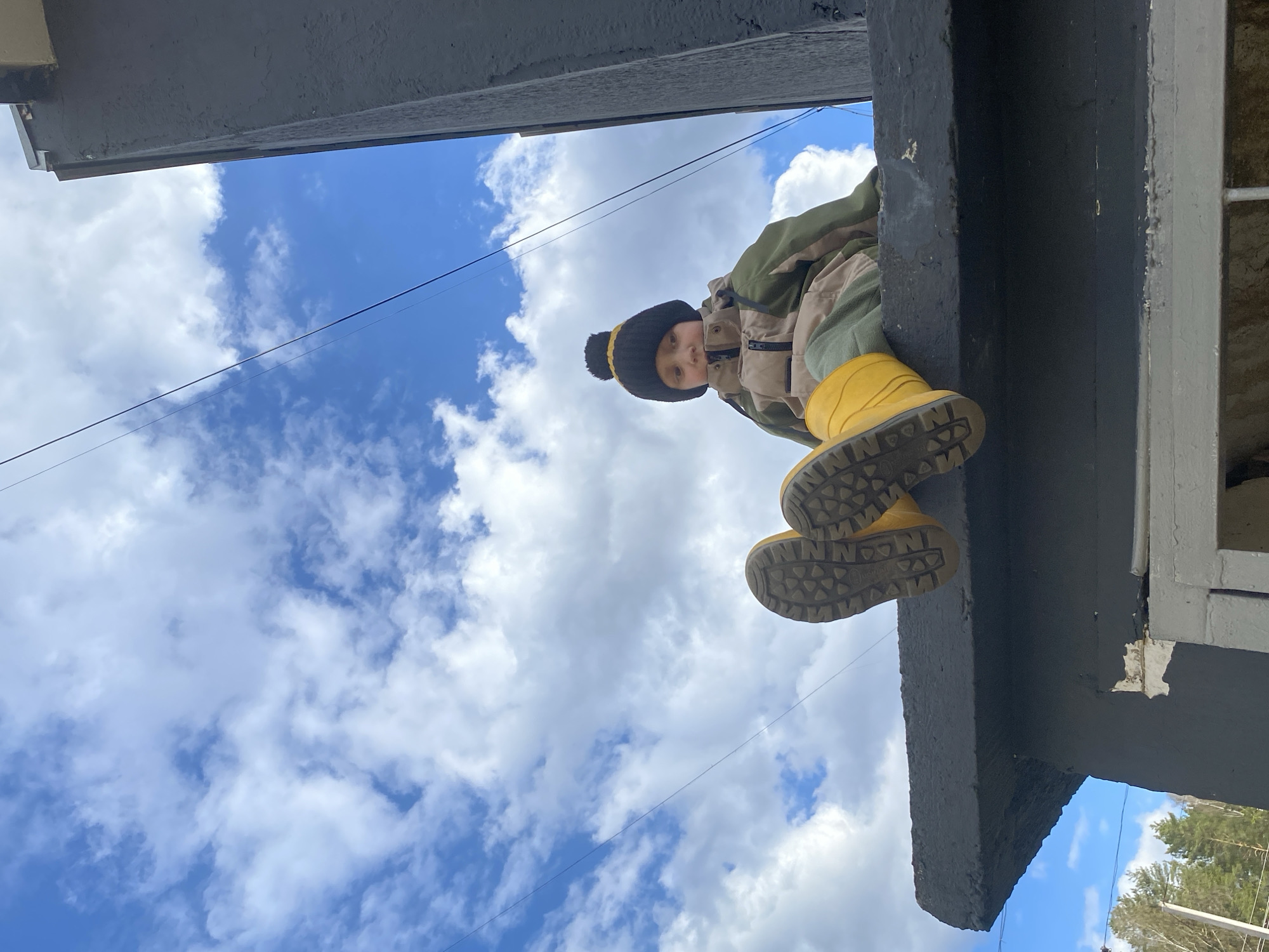 A low-angle shot, taken in the city of Noviy, Russia, captures a child in yellow rain boots, a tan and green jacket, and a black and yellow pom-pom hat, positioned above the camera. The child is standing on a gray-colored ledge, which is attached to a building. The background is a bright blue sky filled with fluffy white clouds. A power line stretches across the sky behind the child. The overall perspective gives the impression that the viewer is looking up at the child from below.