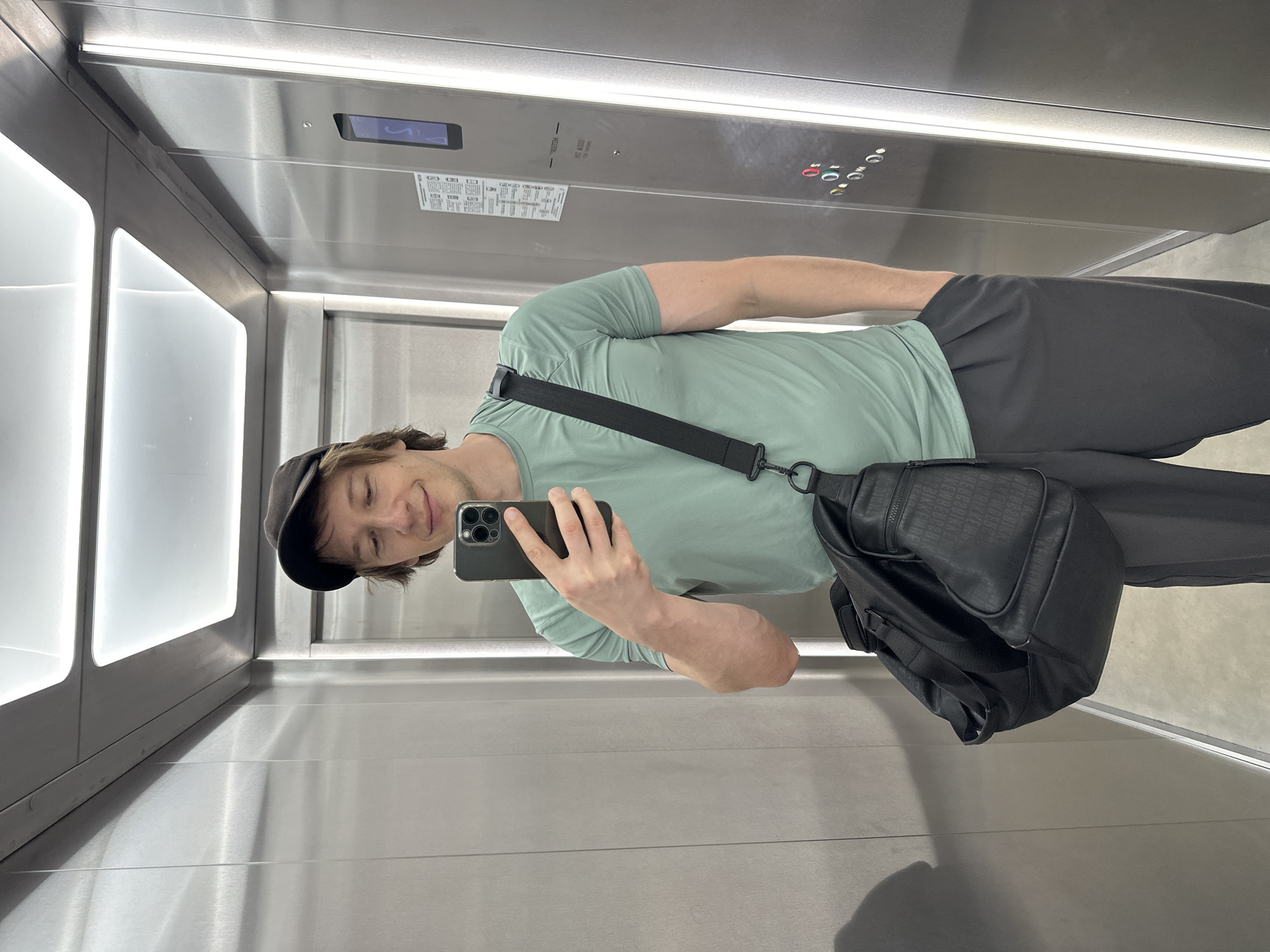 A young man is taking a selfie in an elevator. He is wearing a light green t-shirt, dark pants, and a black baseball cap. He is holding a black smartphone in his right hand and has a black crossbody bag slung over his left shoulder. The elevator appears to be modern, with stainless steel walls and bright LED lighting. On the elevator wall, there is a digital display showing "5", and a panel with buttons labeled with numbers and symbols. There is also a sticker with Russian text and diagrams. The man has a slight smile on his face, suggesting a casual and possibly cheerful mood. The lighting in the elevator is bright, indicating it's likely daytime. The presence of the elevator suggests an indoor setting, possibly a residential building or office complex in Murino, Russia. The overall impression is one of a candid, everyday moment captured in a contemporary urban environment.