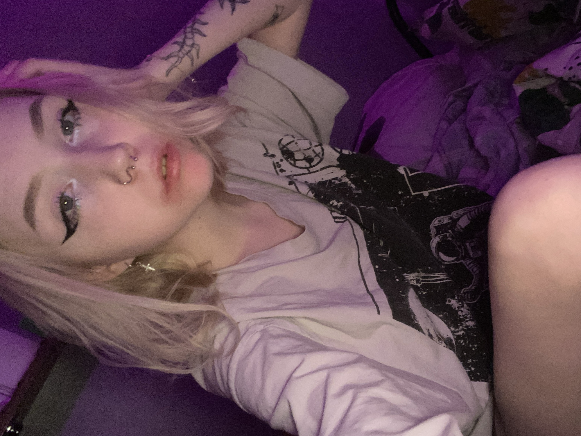 The image presents a close-up, eye-level portrait of a single individual, appearing to be a young person with light-colored (blonde/platinum) hair. The subject is looking directly into the camera. Visible facial features include blue or green eyes accented with black winged eyeliner and white glitter makeup along the lower lash lines. The nose features a twisted barbell septum piercing and two small stud or ring piercings on the nostrils. A small cross-shaped dangle earring is present on the subject's left earlobe. The mouth is slightly open, revealing some upper teeth, with lips appearing pale or subtly colored.
The subject is wearing a light-colored (possibly white or light grey) short-sleeved t-shirt with a dark graphic print on the front, depicting an astronaut figure and abstract elements, possibly related to space themes. The subject's left forearm, visible on the right side of the frame, displays multiple black line-work tattoos of organic or abstract designs. The left hand rests near the subject's head, partially obscured by hair.
The scene is illuminated by strong purple/magenta ambient lighting, which saturates the entire image. The background is indistinct and uniformly dark purple, indicating an indoor setting with minimal visible detail. A dark horizontal line or edge is discernible in the upper left corner of the frame. The specific city of Izhevsk, Russia, or any precise geographic location, cannot be determined from the visual evidence provided in the image.