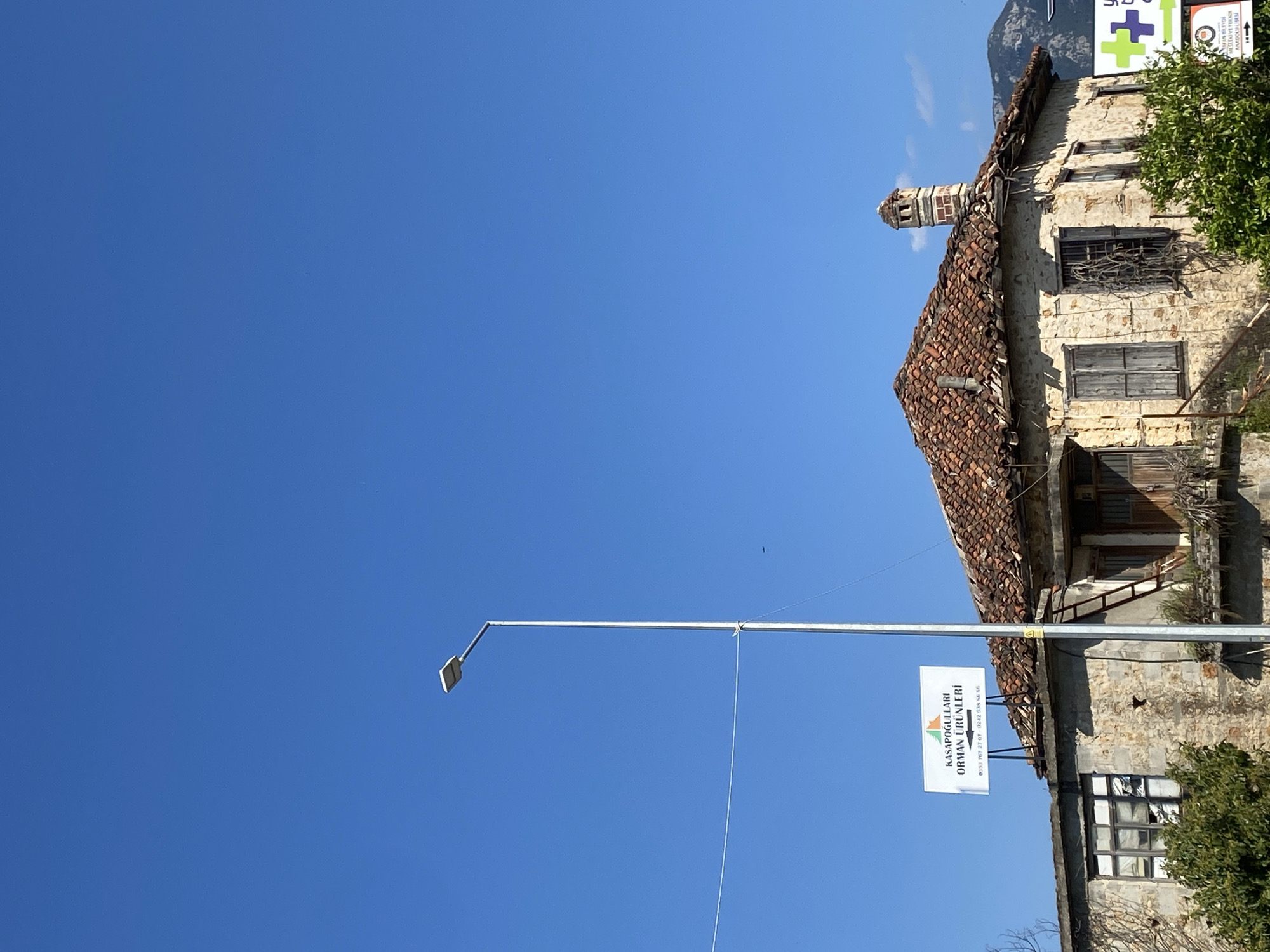 The image displays an old stone building, a modern street light, and a clear blue sky on a bright, sunny day. The dominant feature is the expansive, vibrant blue sky, which fills the upper and left parts of the frame.
On the right side, an aged, two-story stone building stands with a traditional, weathered red-tiled roof and a distinctive tall brick chimney. The building features old wooden window frames, some with shutters, and appears to be in a state of disrepair, with visible stone masonry and some green foliage growing around its structure.
A light grey street light pole extends from the lower left towards the right, with a modern LED light fixture on its left end. A white rectangular sign is suspended from this pole. The sign's text, in Turkish, reads "KASAPOĞULLARI ORMAN ÜRÜNLERİ" (Kasapogullari Forest Products), followed by two phone numbers: "0553 767 27 07" and "0242 538 56 56". In the far upper right corner, another partially visible sign with a green cross symbol is present, displaying "MESLEKİ VE TEKNİK ANADOLU LİSESİ" (Vocational and Technical Anatolian High School).
Distant mountains are faintly visible in the background, specifically on the far right behind the building. The bright sunlight indicates it is daytime. The Turkish text and architectural style provide location cues, consistent with the provided context of Alanya, Turkey. No people or specific activities are depicted in the static scene.