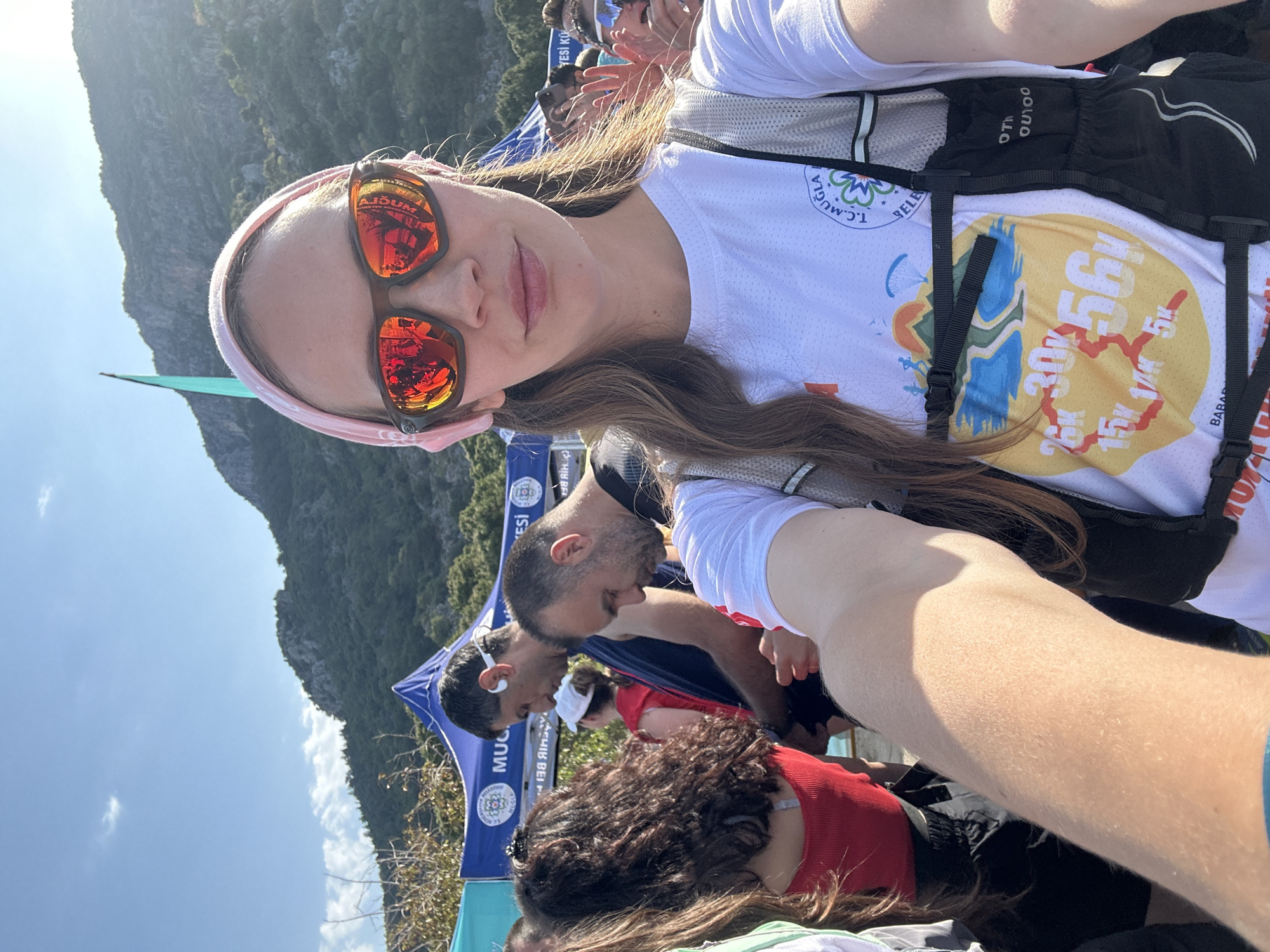 The image depicts a woman taking a selfie outdoors, likely at an event in Fethiye, Turkey. She is wearing sunglasses with red lenses and a pink headband. She has on a white t-shirt with what appears to be a race logo and a black hydration pack. In the background are other people, event tents with the words "MUC" and "EHIB BEI" visible, and a view of mountainous terrain. The scene appears to be a gathering or event related to a running race.