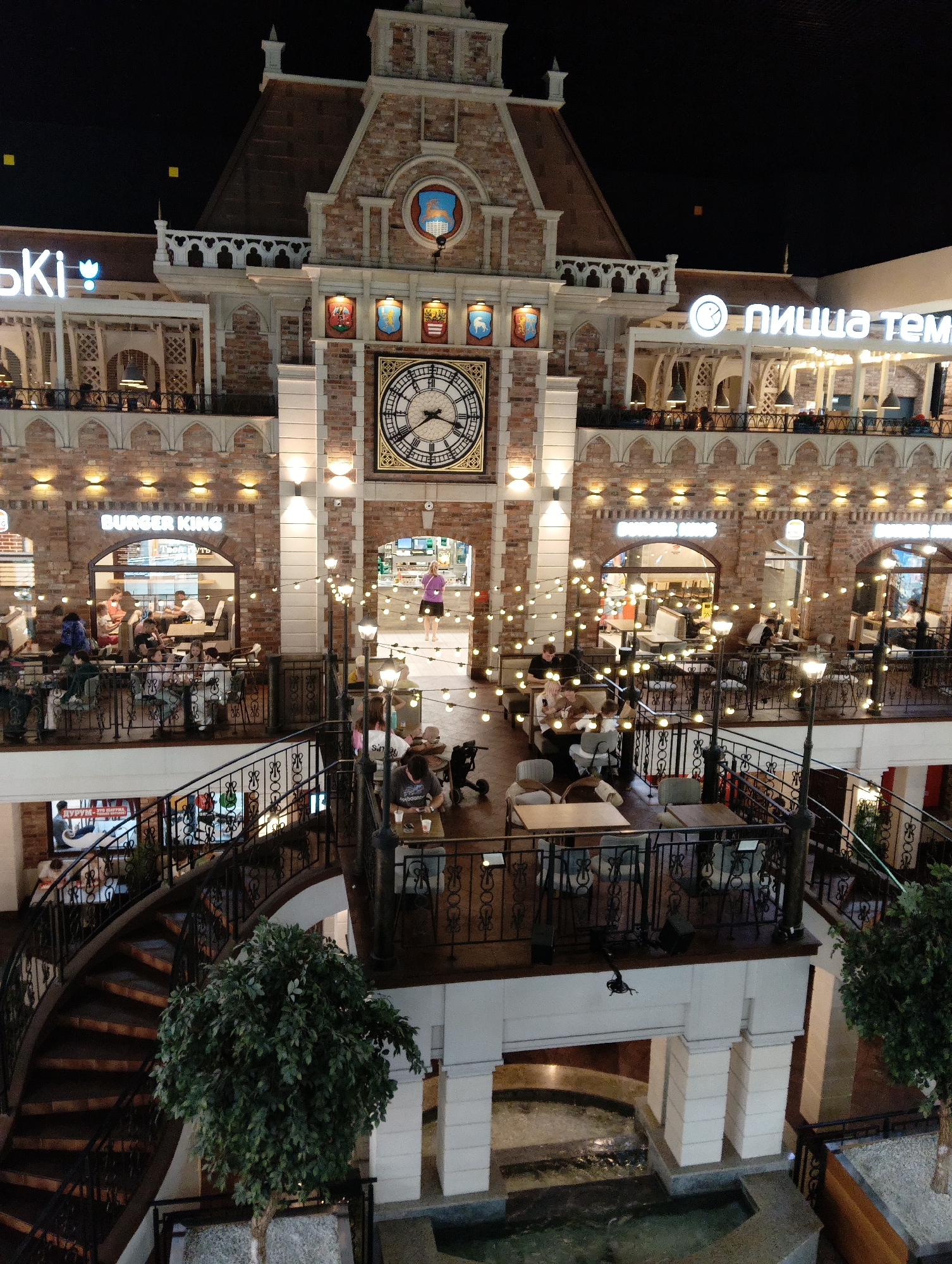 This is an indoor, multi-level shopping mall or entertainment center. The architecture features brick walls, arched windows, and decorative balconies with wrought-iron railings. A large clock face is prominent on the facade. String lights are draped throughout the scene, illuminating several dining areas. People are seated at tables in restaurants and cafes on different levels. A spiral staircase connects the upper and lower levels. There are also decorative trees and a water feature visible in the lower foreground. The overall ambiance suggests a lively commercial space, possibly a food court or a section of a larger complex. Based on the Cyrillic text visible on signs (e.g., "Burger King", "пицца темп"), and the specific architectural style, this location is likely in Hrodna, Belarus.