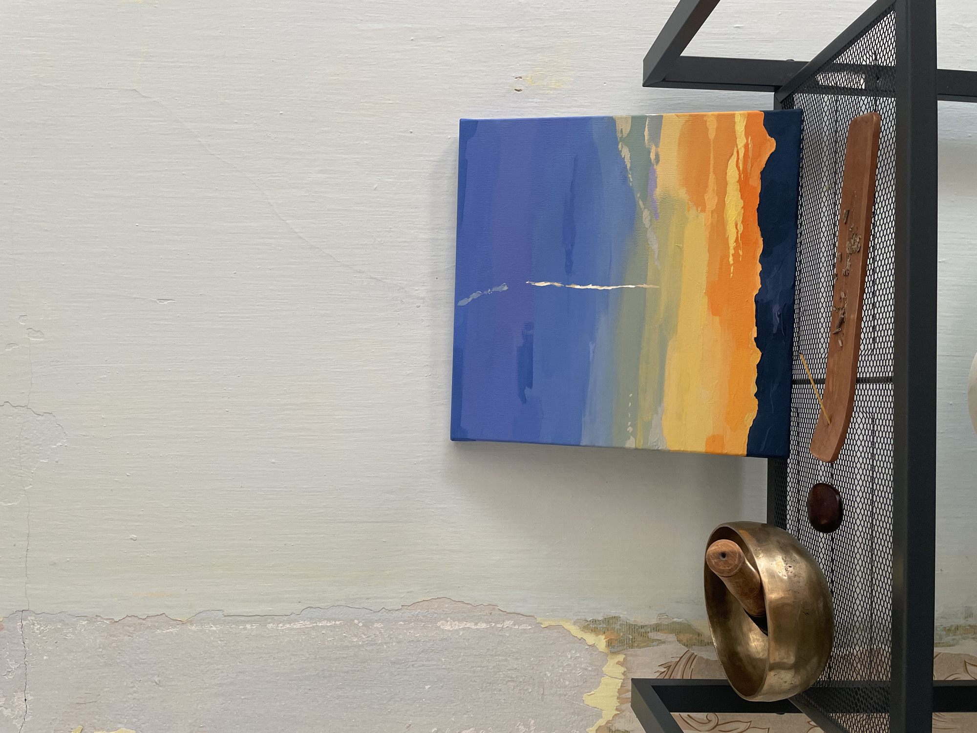 A small, square painting of a sunset hangs on a textured, off-white wall, slightly left of center. The painting depicts a gradient of colors, starting with a deep blue sky at the top, transitioning to shades of orange and yellow at the horizon, with a dark blue silhouette of mountains at the very bottom. A thin, vertical white streak, possibly a contrail, is visible in the upper part of the sky. To the left of the painting, on a black metal shelf, sits a brass singing bowl containing a wooden mallet. Next to the bowl, there is a dark, smooth stone and a wooden incense holder with partially burned incense sticks. The shelf is composed of a mesh grid. The wall behind the shelf shows signs of wear and tear, with peeling paint and visible cracks, particularly on the left side. The overall scene suggests an indoor setting, possibly a room with an artistic or meditative theme. The image does not contain any people or depict any specific urban location within Kopeysk, Russia.