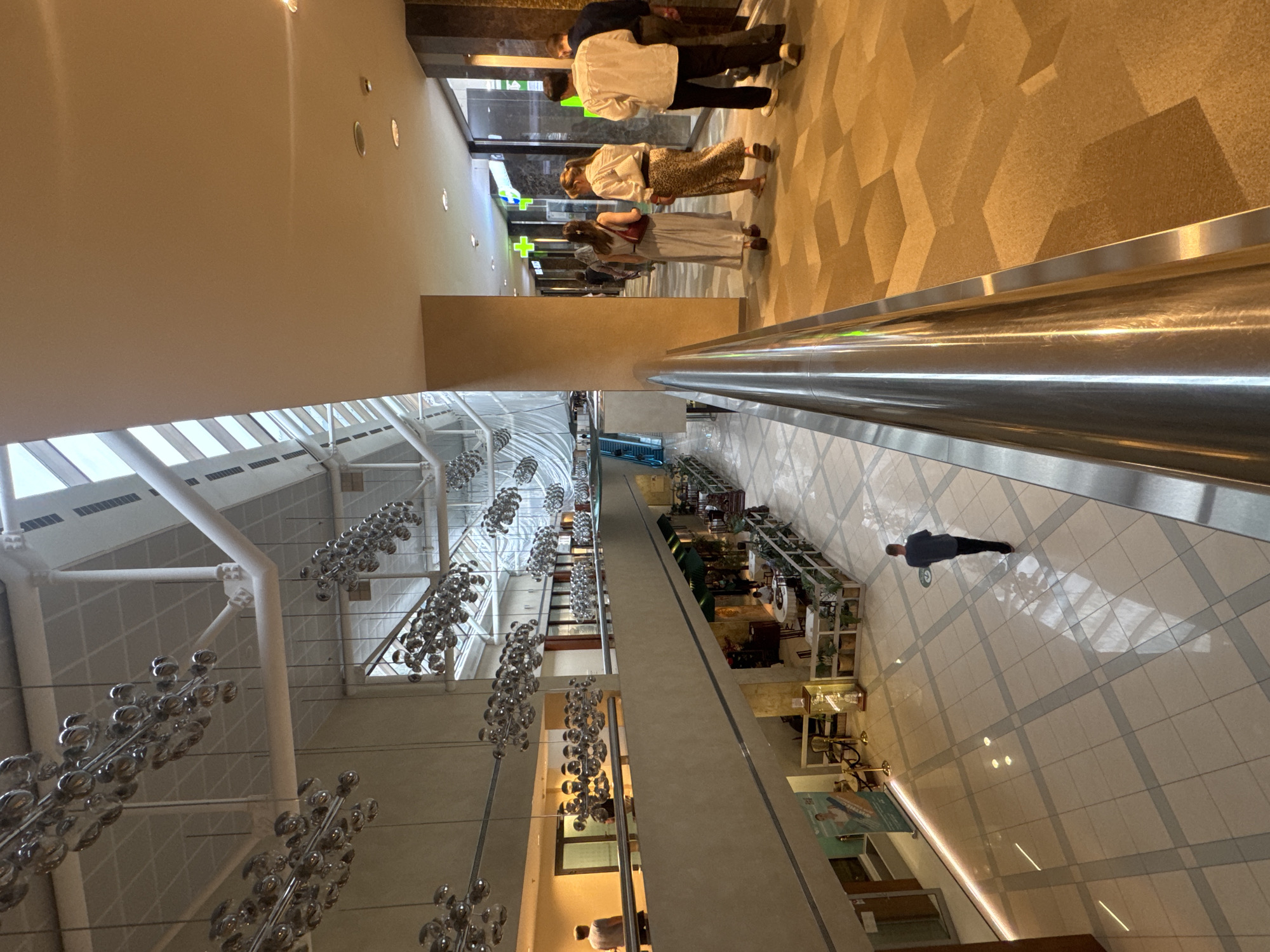 This is an interior shot of an airport in Moscow, Russia. The scene is from an elevated position. The interior space features a high ceiling with a steel frame and large chandeliers of metallic orbs suspended from the ceiling. A group of people are walking through a terminal near storefronts and shops. One person is walking away from the camera in the middle of the tile floor. The scene shows a corridor with shops on one side, and another terminal leading to a pharmacy in the distance.