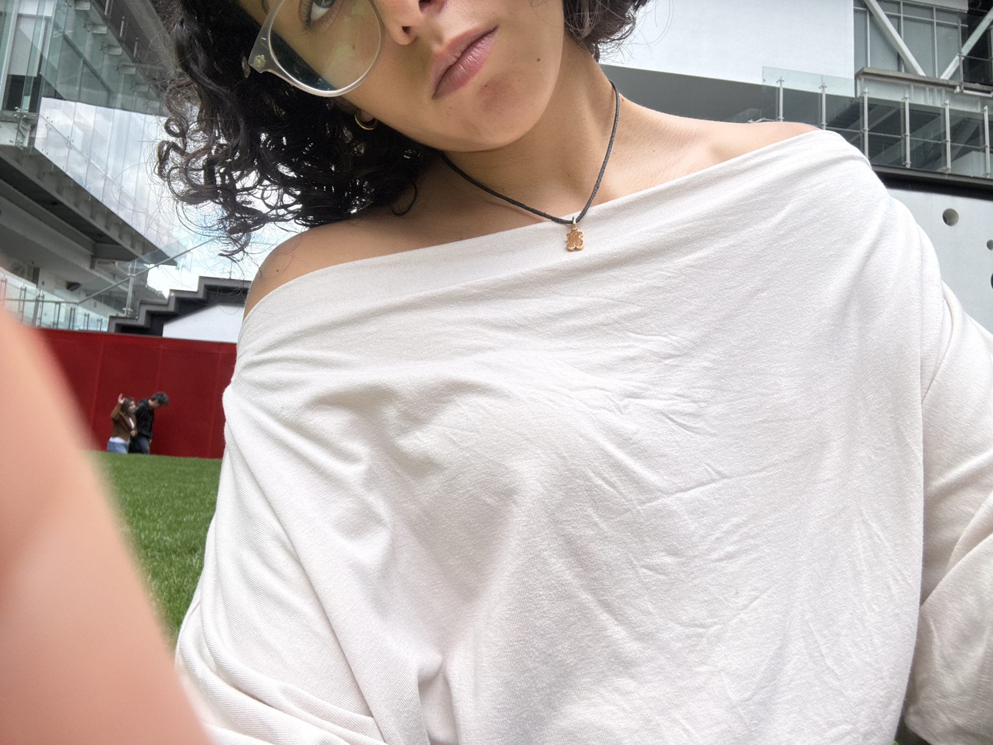 The image is a selfie of a person with curly dark hair, wearing round glasses and a white off-the-shoulder top with a black necklace with a gold teddy bear pendant. The person has neutral lip color.
In the background, there is a modern building with glass walls and metal structures. There is also a patch of green grass.
In the distance, behind a red wall, two people are visible. One is kneeling and the other is standing.
Based on the architecture and the presence of a grass area near a modern building, it is plausible the location is in Mexico City, Mexico, as it is known for its blend of modern architecture and green spaces.