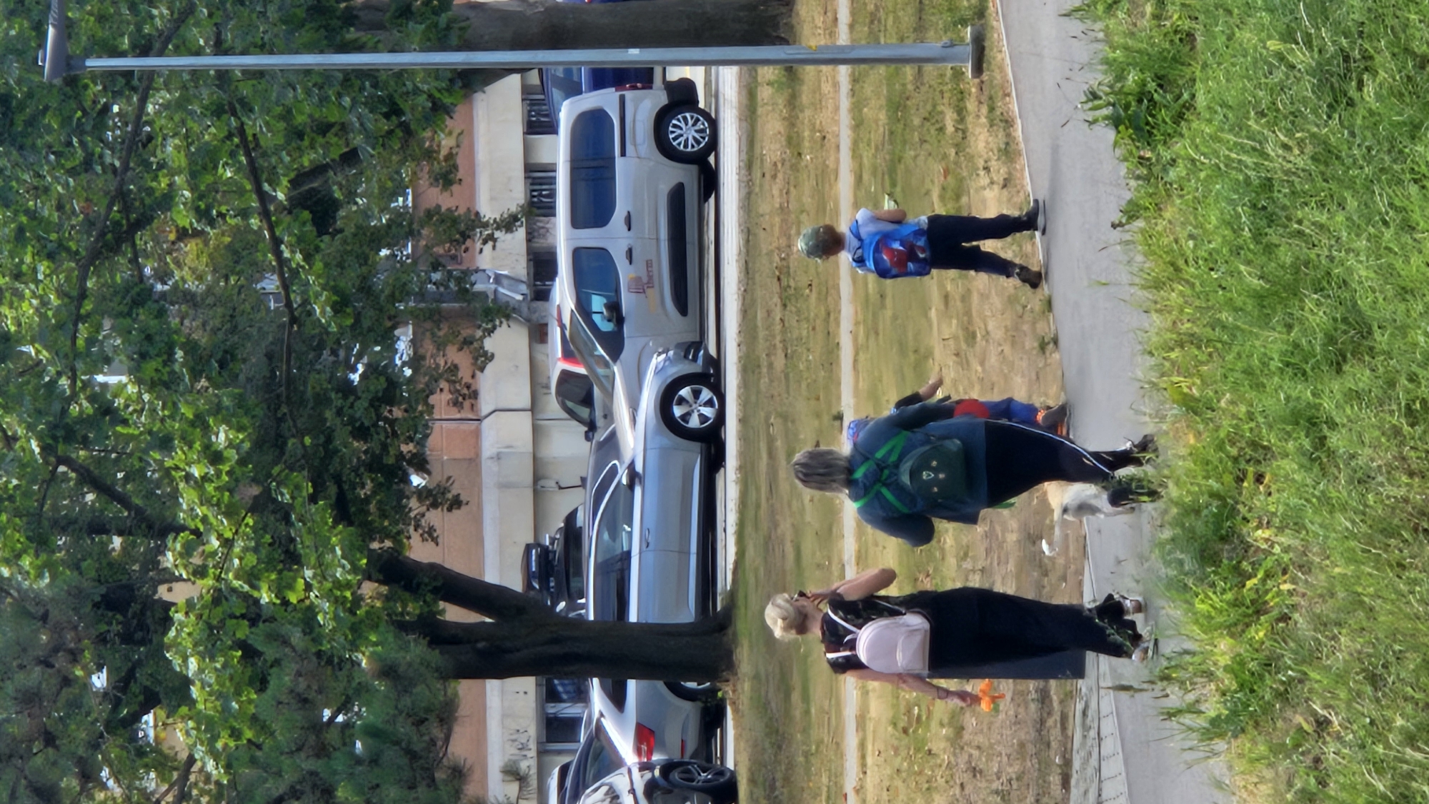 In Prešov, Slovakia, a group of people walks along a paved path in a grassy park on a sunny day. Two women, each wearing a backpack, are in the foreground. The woman on the left, with blonde hair, is wearing a black dress and carrying an orange object. The woman on the right, with dark hair, is wearing a blue backpack and black pants. Behind them, two children walk. One child, wearing a blue backpack with a spider-man design, is ahead of the other child. A light-colored dog on a leash is walking with the woman on the right. In the background, cars are parked on a street, with a row of buildings visible behind them. Trees with lush green leaves provide shade over the park.