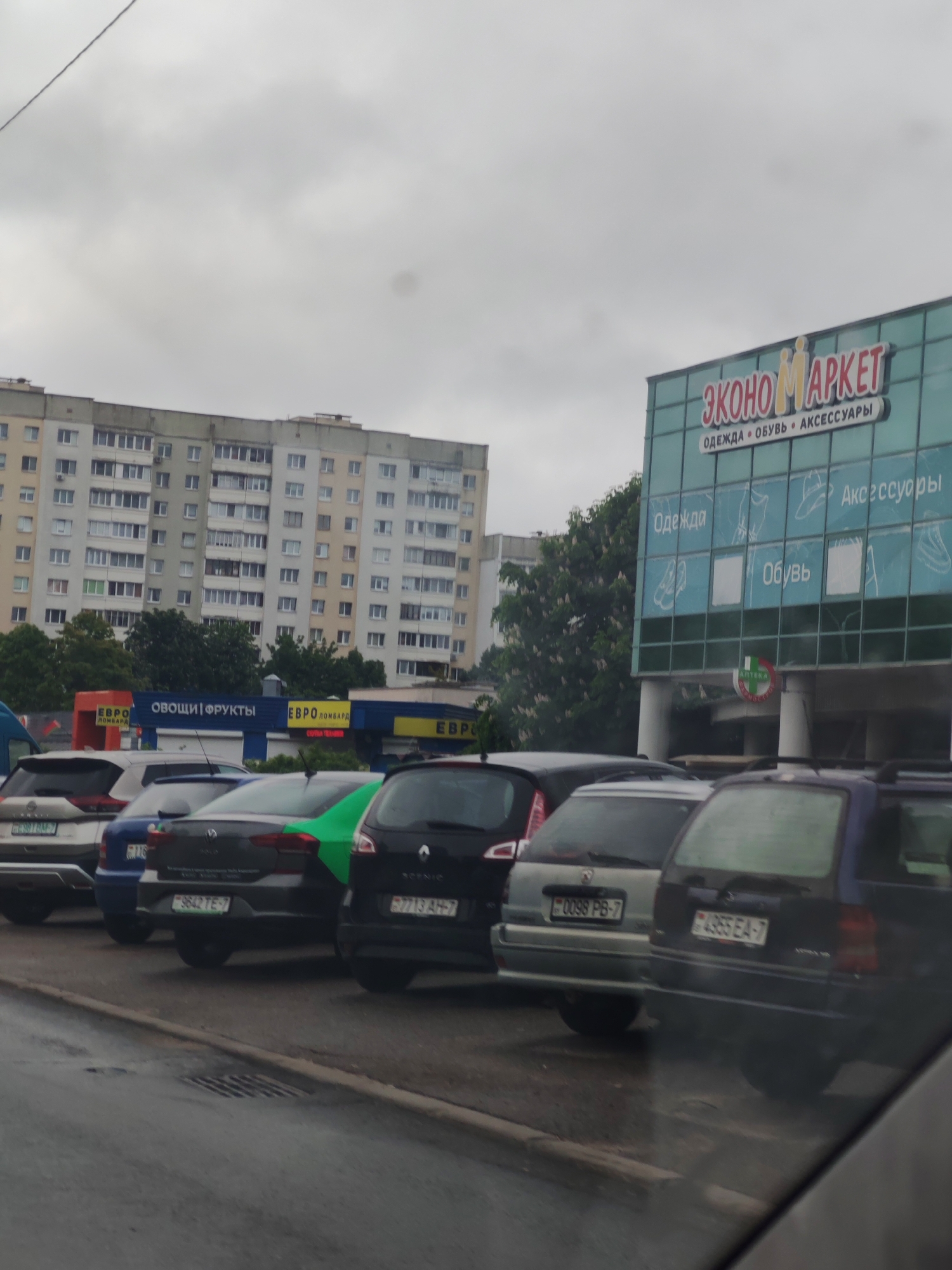 Here is a detailed description of the image:
The image depicts a row of parked cars in front of a building on an overcast day. The building has a large sign reading "ЭКОНОМАРКЕТ" (ECONOMARKET) with smaller text below indicating "ОДЕЖДА • ОБУВЬ • АКСЕССУАРЫ" (CLOTHES • SHOES • ACCESSORIES). Pictorial representations of clothing, shoes, and accessories are displayed on the glass façade. Below the Economarket sign, there's a smaller circular sign with "AПТЕКА" (Pharmacy) visible.
Adjacent to the Economarket, there are signs related to "ЕВРО ЛОМБАРД" (EURO PAWNSHOP) and "ОВОЩИ | ФРУКТЫ" (VEGETABLES | FRUITS). A large apartment building is visible in the background.
The row of parked cars includes vehicles with license plates displaying the format "#### **-**" suggesting the location may be in Belarus. The visible license plates read "9642 TE-7", "7713 AH-7", "0098 PB-7", and "4955 EA-7".
The road surface appears wet, suggesting recent rainfall. The photo is taken from inside a vehicle, as evidenced by a partial view of the dashboard or window frame.