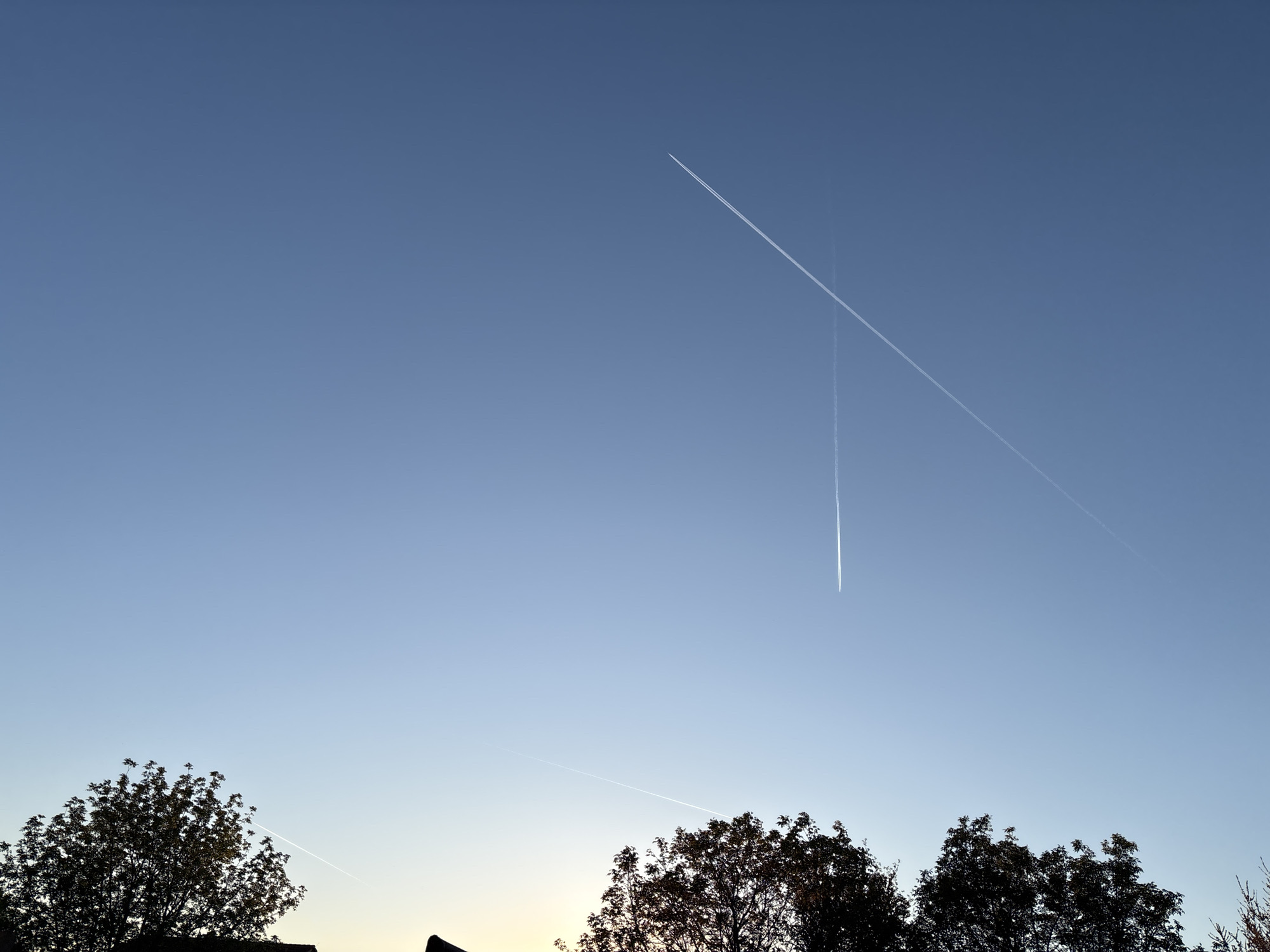 The image depicts a blue sky at dusk with multiple contrails from airplanes visible. The contrails intersect at one point. Below, there is a line of trees in silhouette. A portion of a rooftop is also visible. While the location cannot be verified from the image itself, the prompt specifies that it should be considered Wiener Neustadt, Austria.