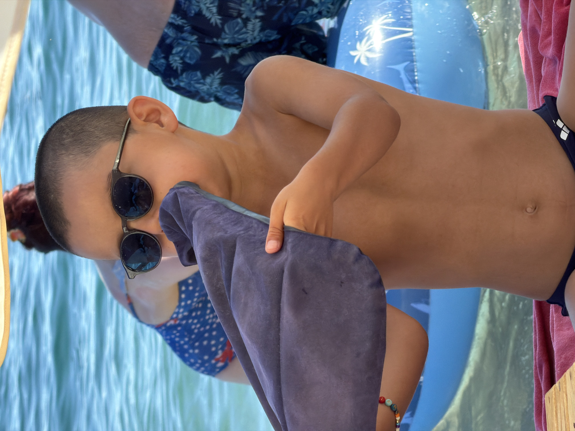 The image captures a close-up, vertical shot of a young person at the beach in Budva, Montenegro. The person, wearing dark sunglasses and a blue and white polka dot swimsuit, is holding a dark purple towel, partially obscuring their face. They have a shaved head and appear to be topless. Behind them, a bright blue inflatable ring with palm tree graphics is visible, suggesting a playful and relaxed atmosphere.
The setting is clearly a beach or poolside, indicated by the presence of water and the inflatable ring. The water is a light blue, reflecting sunlight, and ripples are visible on its surface. To the left, a hint of a wooden surface suggests a boardwalk or pier. A person wearing dark blue swim trunks with a floral pattern is partially visible in the background, adding to the sense of a family or group outing.
The lighting suggests it is daytime, likely sunny, given the bright reflections on the water. The overall mood is casual and summery. The focus is on the person and their interaction with the towel, with the background providing context. No visible text is present in the image.
