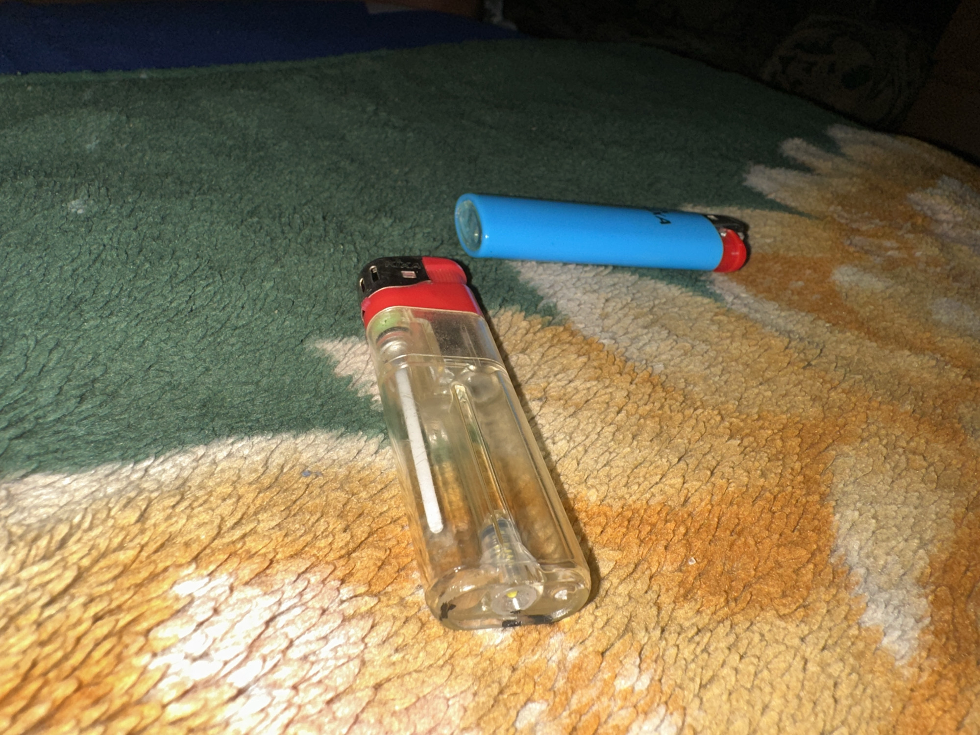 The image displays two lighters resting on a soft, textured surface, likely a blanket or rug, in an indoor setting.
In the foreground, a transparent lighter with a red top and black firing mechanism is visible. It features an integrated LED light at its base, suggesting a dual function as a small flashlight. Further back and to the right, a second, opaque blue lighter is present, also with a red firing mechanism. Partial text "KA" is discernable on the side of the blue lighter.
The underlying surface exhibits a varied pattern and texture. The area around the lighters is predominantly orange and cream, featuring distinct, slightly raised fibrous patterns. This transitions into a solid dark green section in the middle and upper-left background. A darker, possibly blue, indistinct area is visible in the far upper background.
The lighting is dim, creating a subdued atmosphere and highlighting the textures of the fabric. There is no specific activity or event taking place; the lighters are static. No people are visible, and no emotions are conveyed. The time of day is not explicitly clear but the dim lighting might suggest evening or night. While the location context is Jos, Nigeria, the image itself does not contain specific visual cues to confirm this.