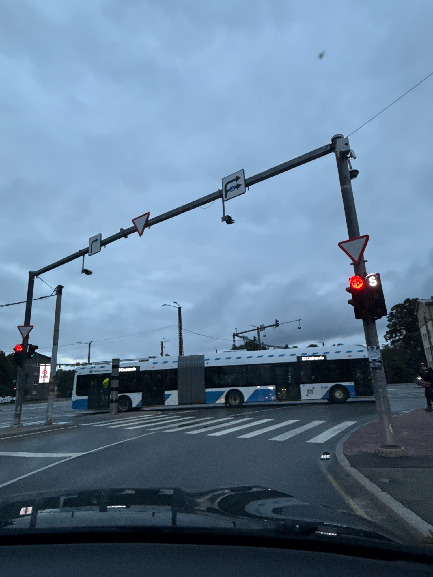 The image depicts a street scene in Tallinn, Estonia, under an overcast sky. In the foreground, a vehicle's dashboard is visible. In the mid-ground, a long articulated bus with blue and white markings is stopped at a pedestrian crossing. The bus displays "Tallinna Transport" on its side. Traffic lights are present, with the one on the right showing a red signal. Road signs, including a yield sign and directional signs, are mounted on overhead poles. In the background, trees and buildings are visible, along with lampposts and other street infrastructure. A few people are present on the sidewalk to the right. The overall atmosphere is dim, suggesting it is either early morning or late evening.