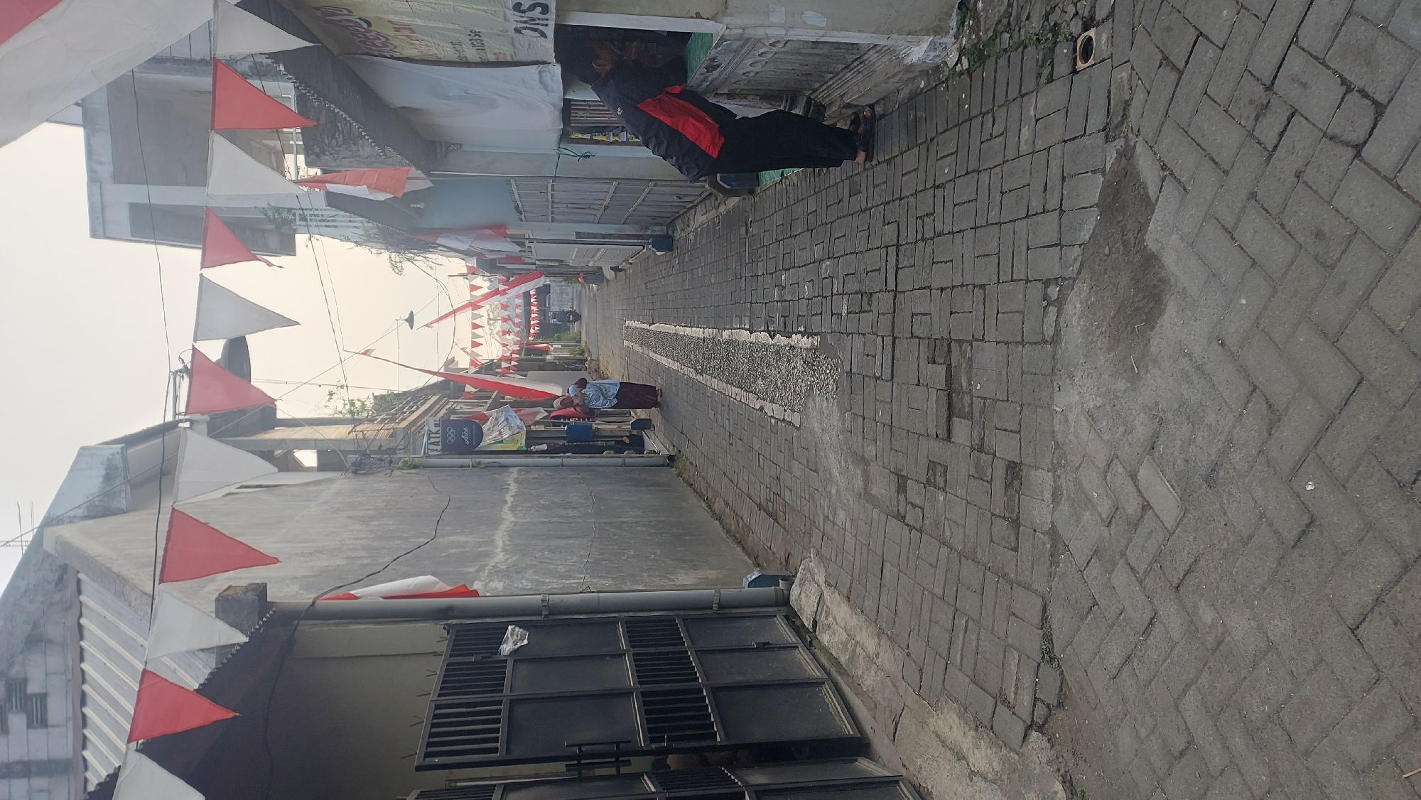 An alleyway in Malang, Indonesia, is decorated with red and white triangular flags, suggesting a national celebration. The alley is paved with interlocking bricks and flanked by narrow houses. In the mid-ground, a woman in a headscarf and long skirt walks away from the camera. Further down the alley, a person in a black jacket with red accents leans into a window of a building on the right, seemingly interacting with someone inside. The sky is overcast and hazy, indicating a possible early morning or late afternoon setting. A motorcycle is parked in the distance.