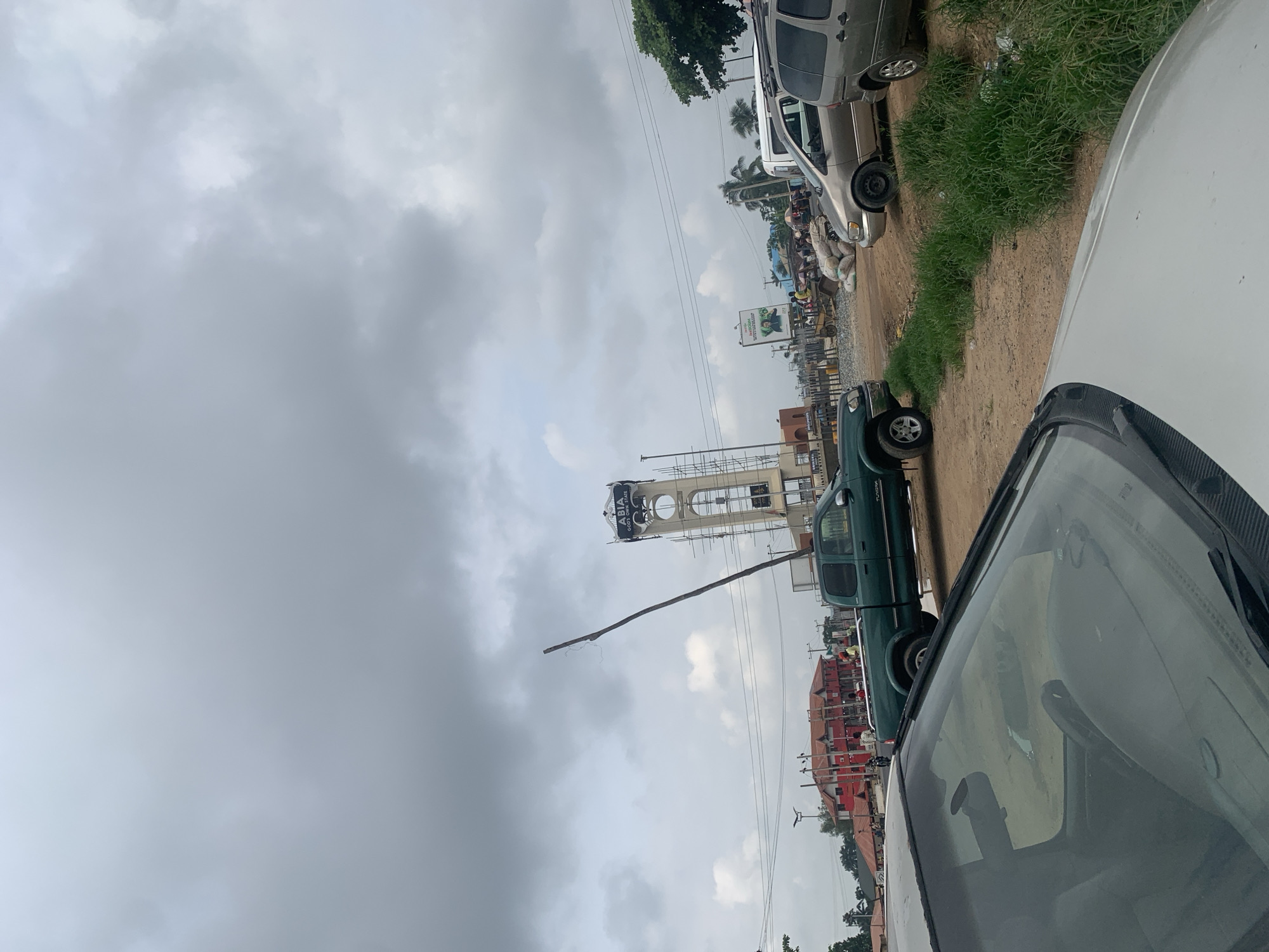 The image captures a view of a roadside scene in Mbara Akuma, Nigeria, under a cloudy sky. A prominent clock tower, bearing the inscription "ABIA GOD'S OWN STATE," stands in the background, partially obscured by scaffolding. In the foreground, a silver car is parked, with its windshield and dashboard visible. To the right of the car, a green pickup truck is parked on a dirt road. Several other vehicles, including vans and SUVs, are also present, along with a scattering of bags of what appears to be cement or sand. A few individuals are visible near the vehicles and in the distance, suggesting a lively commercial or transit area. The environment appears dusty with patches of overgrown grass.