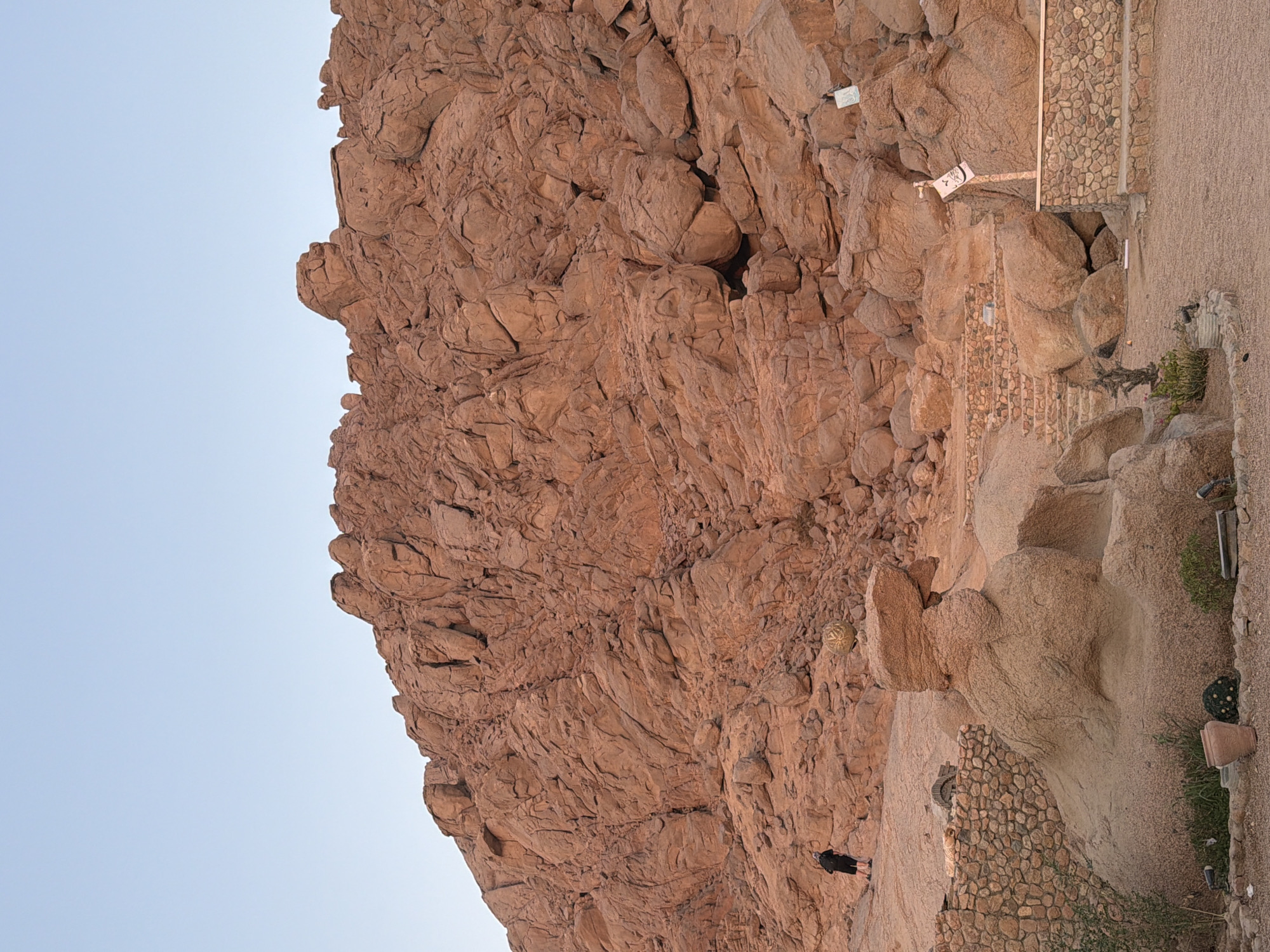 In Sharm El Sheikh, Egypt, a person wearing a black jacket and shorts walks on a dusty path towards a massive, rugged mountain composed of reddish-brown rock. The mountain dominates the landscape, with its textured surface showing deep fissures and large boulders. In the foreground, a unique rock formation resembling a profile view of a face or animal head is visible. Adjacent to this formation is a decorative stone wall and various potted plants, including a large ceramic pot. Steps made of stone lead upwards into the rocky terrain. Signs with Arabic inscriptions are placed on poles near the rock face. The sky above is a clear, pale blue, indicating daytime.