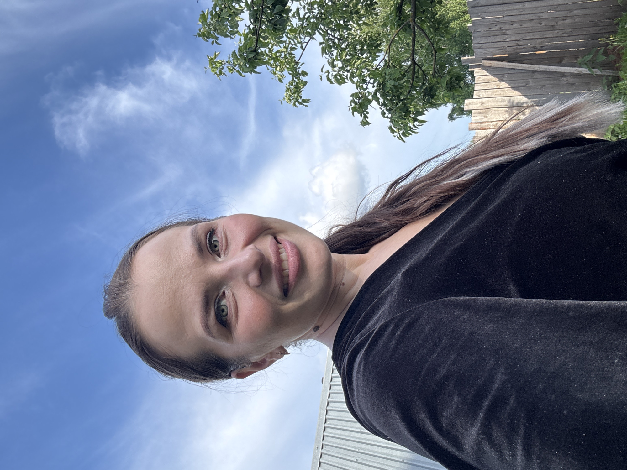This media file features a close-up, low-angle selfie of a smiling woman outdoors in Rusalevka, Russia. The woman, with long, dark brown hair with lighter streaks and green eyes, is looking towards the camera with a broad smile, her cheeks slightly flushed. She is wearing a black top, possibly made of velvet or a soft, textured fabric.
The image is oriented with the woman's face at the bottom and center, looking upwards. The background primarily consists of a bright, clear blue sky with scattered white, wispy clouds, indicating daytime and good weather. To the upper right, green tree branches and leaves are visible. On the far right, a weathered wooden fence made of horizontal planks forms part of the background, and beneath it, a grey corrugated or paneled surface, possibly part of a shed or building, can be seen. The natural lighting suggests it is sunny. No visible text is present in the image. The overall impression is one of contentment in an outdoor, likely rural or suburban, setting.