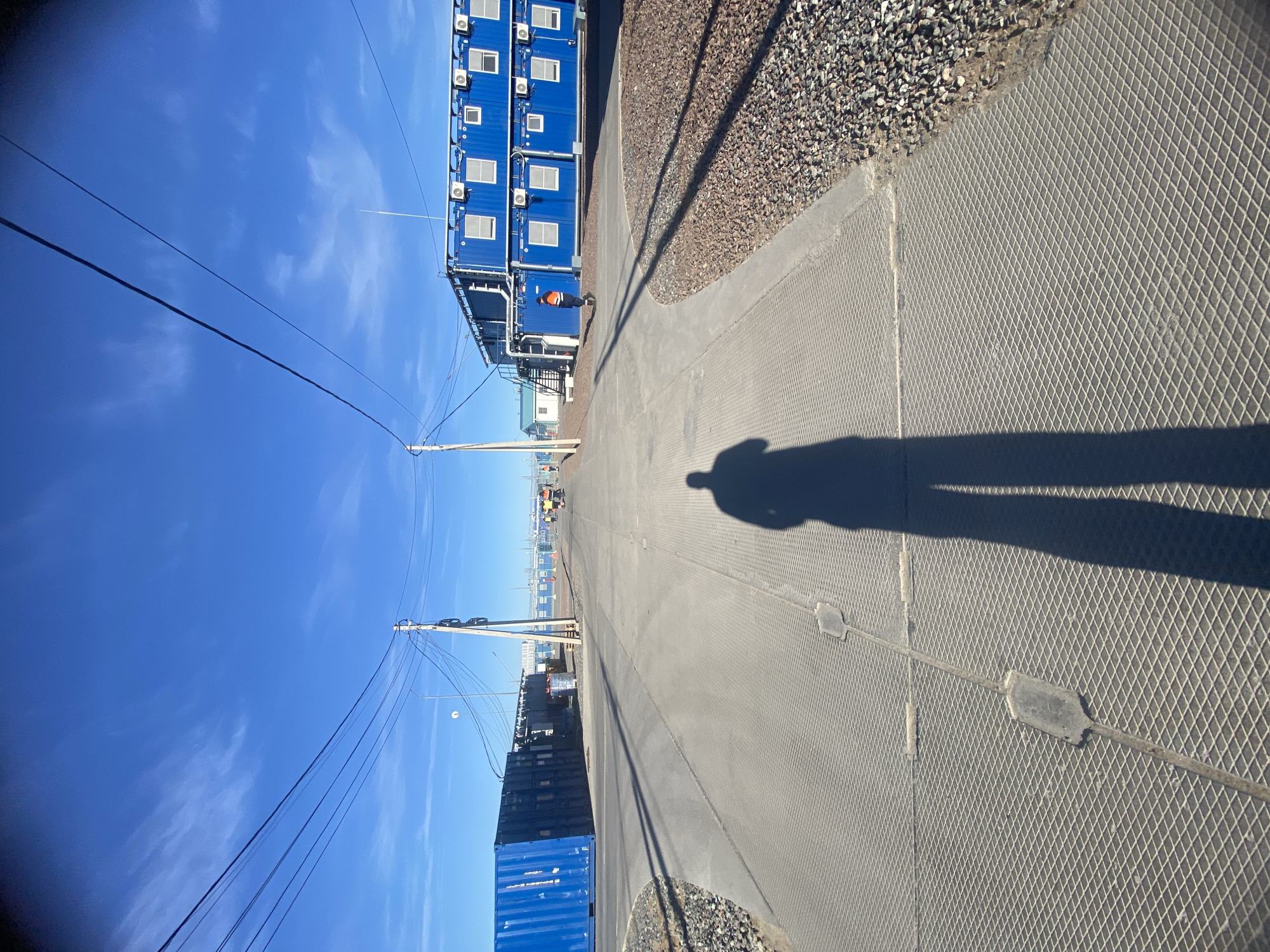 An outdoor, industrial or construction site is depicted under a clear, bright blue sky with sparse, wispy clouds. The image is oriented vertically, with the sky at the top.
In the foreground, a prominent shadow of a person, likely the photographer, stretches across a textured ground surface on the right. This ground surface, which forms a path or walkway, features a diamond grid pattern. To the left of this path, there's a strip of dark gray gravel or crushed stone.
On the left side of the scene, a row of blue, multi-story modular buildings, possibly converted shipping containers, stands tall. These buildings feature multiple windows and external air conditioning units. A person wearing an orange safety vest is visible near the base of these structures. Further down the path, another large blue shipping container is seen, with partial white text visible including "EVERGREEN" and "NYK Line." Utility poles with numerous power lines run along the left side of the path, extending into the distance.
The path, with its distinctive grid pattern, stretches into the background, where more industrial structures and what appears to be heavy machinery, possibly a yellow excavator or loader, are faintly visible. The long, distinct shadows cast by the person and other elements suggest that the sun is relatively low in the sky, indicating either morning or late afternoon. The overall setting appears to be a functional, operational area, consistent with an industrial or logistical facility in a location like Koskolovo, Russia.
