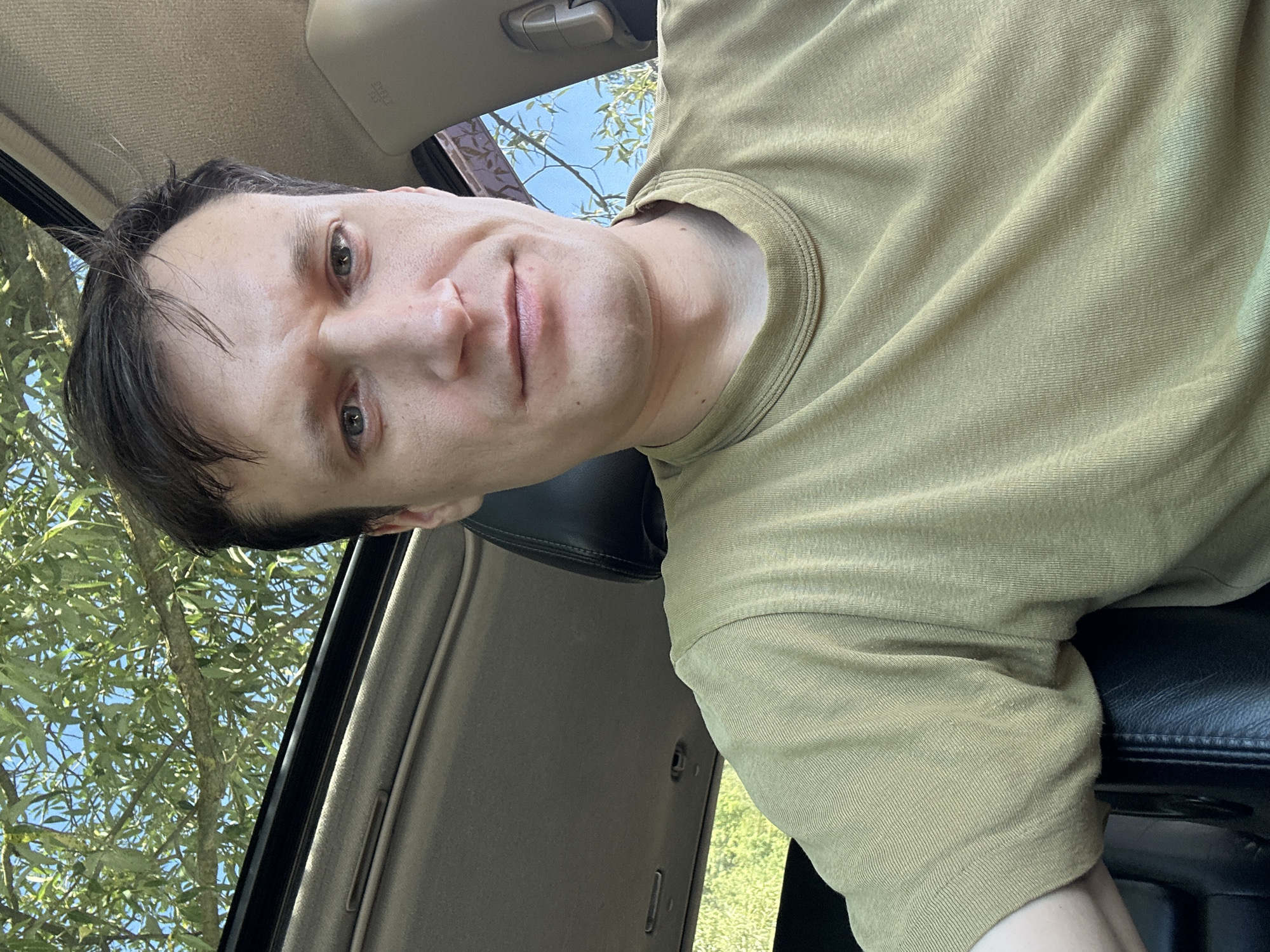 A selfie taken from inside a vehicle shows a Caucasian man with short, dark brown hair and light-colored eyes. He is wearing a short-sleeved, olive-green t-shirt. The man is positioned in the driver's seat, with the sunroof open above him, revealing green foliage and a blue sky. The interior of the car is visible, including the dashboard and headrest. The man's expression is neutral. The scene appears to be outdoors in a rural or suburban setting, possibly in Makiv, Ukraine, given the context.