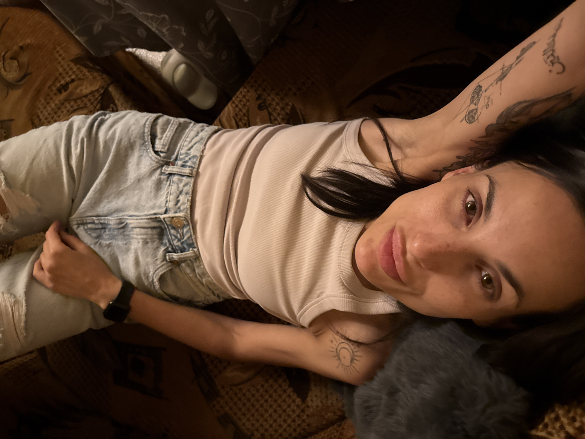 A young woman is lying down, likely on a bed or couch, indoors. She is wearing light blue denim shorts, a light-colored tank top, and a black smartwatch on her left wrist. She has tattoos on both arms and collar bone. Her hair is dark and her eyes are light brown. The background appears to be a patterned brown and beige fabric, possibly a bedspread or couch. The location in the city of Pleven, Bulgaria is undeterminable.