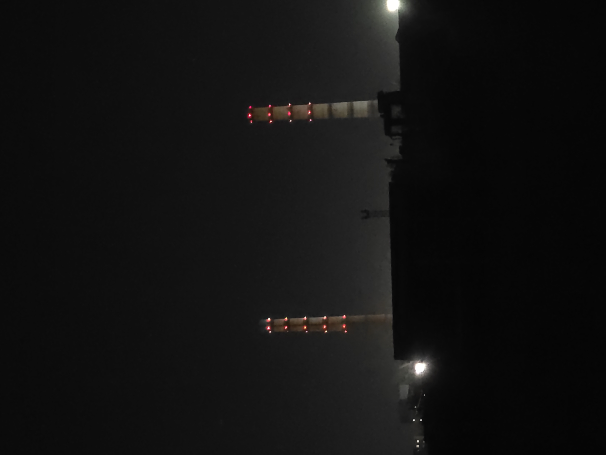 The image shows a night view of the Ust-Kamenogorsk Thermal Power Plant in Oskemen, Kazakhstan. Two tall, industrial chimneys with red lights illuminating their upper sections are prominently visible against the dark sky. The chimneys are cylindrical and appear to be made of concrete or brick. Below the chimneys, a dark silhouette of the power plant's structure is visible, along with some dimly lit lights from other parts of the facility and what appears to be a large, dark building or wall in the foreground. The overall environment is industrial and appears to be a clear night. No people or specific actions are depicted.