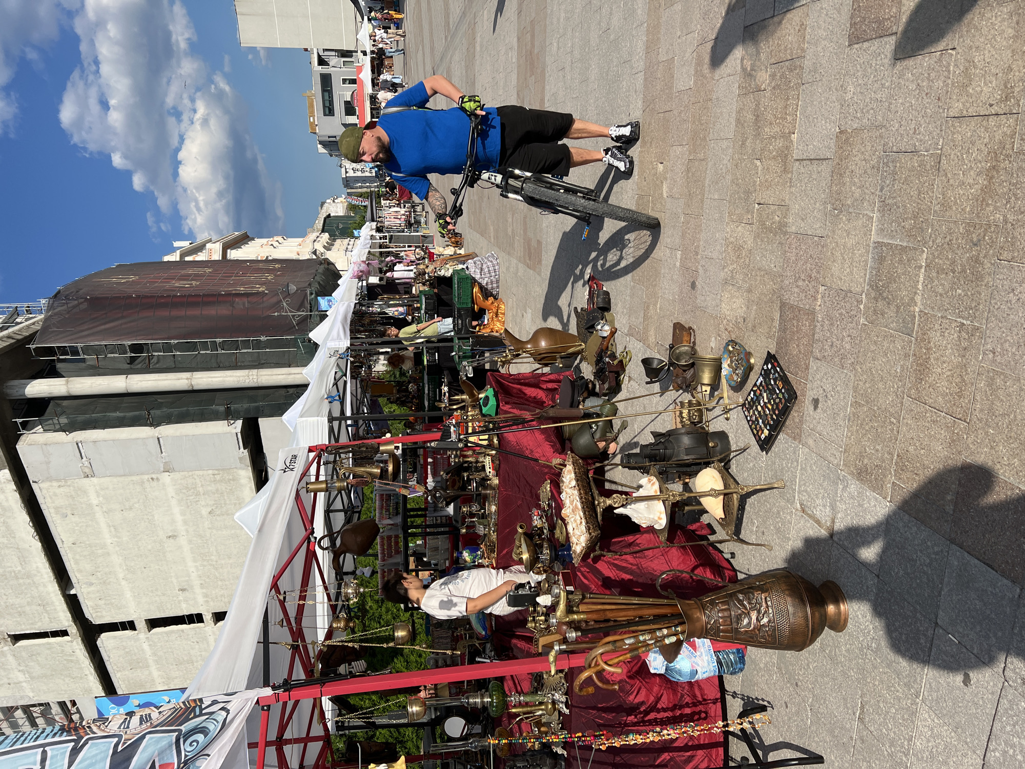 This image depicts an outdoor flea market in Constanța, Romania, likely on a sunny day with a clear blue sky and scattered clouds. In the foreground, a man wearing a blue t-shirt, black shorts, a green cap, and athletic shoes is standing with a bicycle. He has tattoos on his right arm and appears to be looking at the merchandise.
To the left of the man, a market stall is set up under a white tent with a red frame. The stall is draped with a burgundy tablecloth and displays a variety of antique items, including brass objects, decorative vases, walking sticks, and possibly some jewelry. A young person in a white t-shirt and shorts is standing behind the stall, looking at the items.
Further into the background, more market stalls are visible, along with people browsing and walking. Buildings under construction or renovation are present, characterized by concrete structures and scaffolding, suggesting ongoing urban development. The ground is paved with stone tiles. The overall scene conveys a lively atmosphere of an outdoor market.